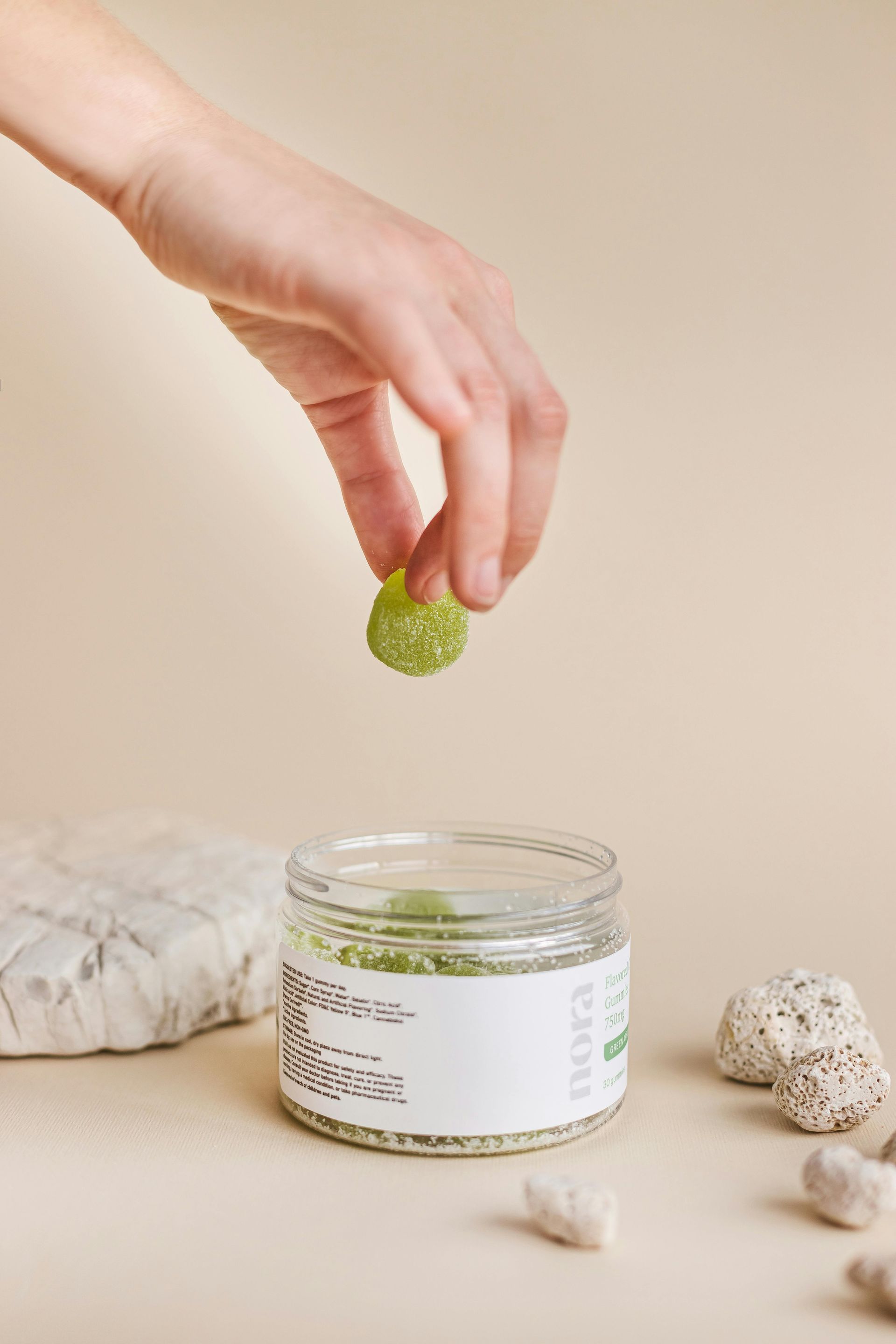 Hand reaching into a jar of green gummy candies, holding one. Beige background, with rocks.