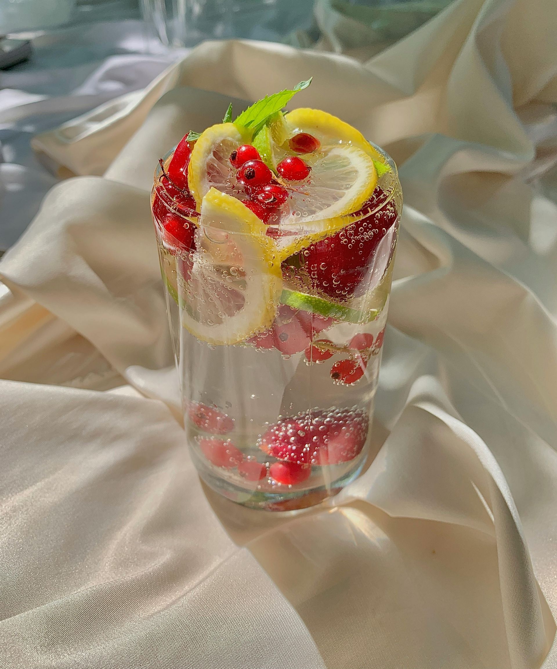 Glass of sparkling water with lemon slices, red berries, and mint, on a beige fabric.