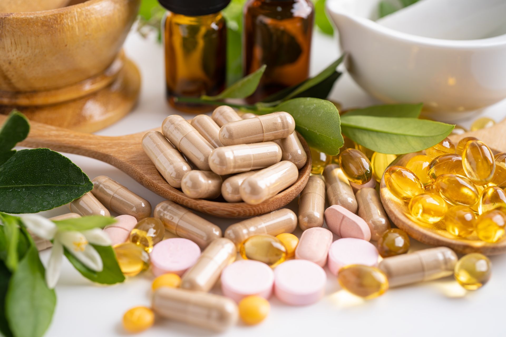 Assortment of pills, capsules, and oils arranged with greenery on a white surface, suggesting natural remedies.