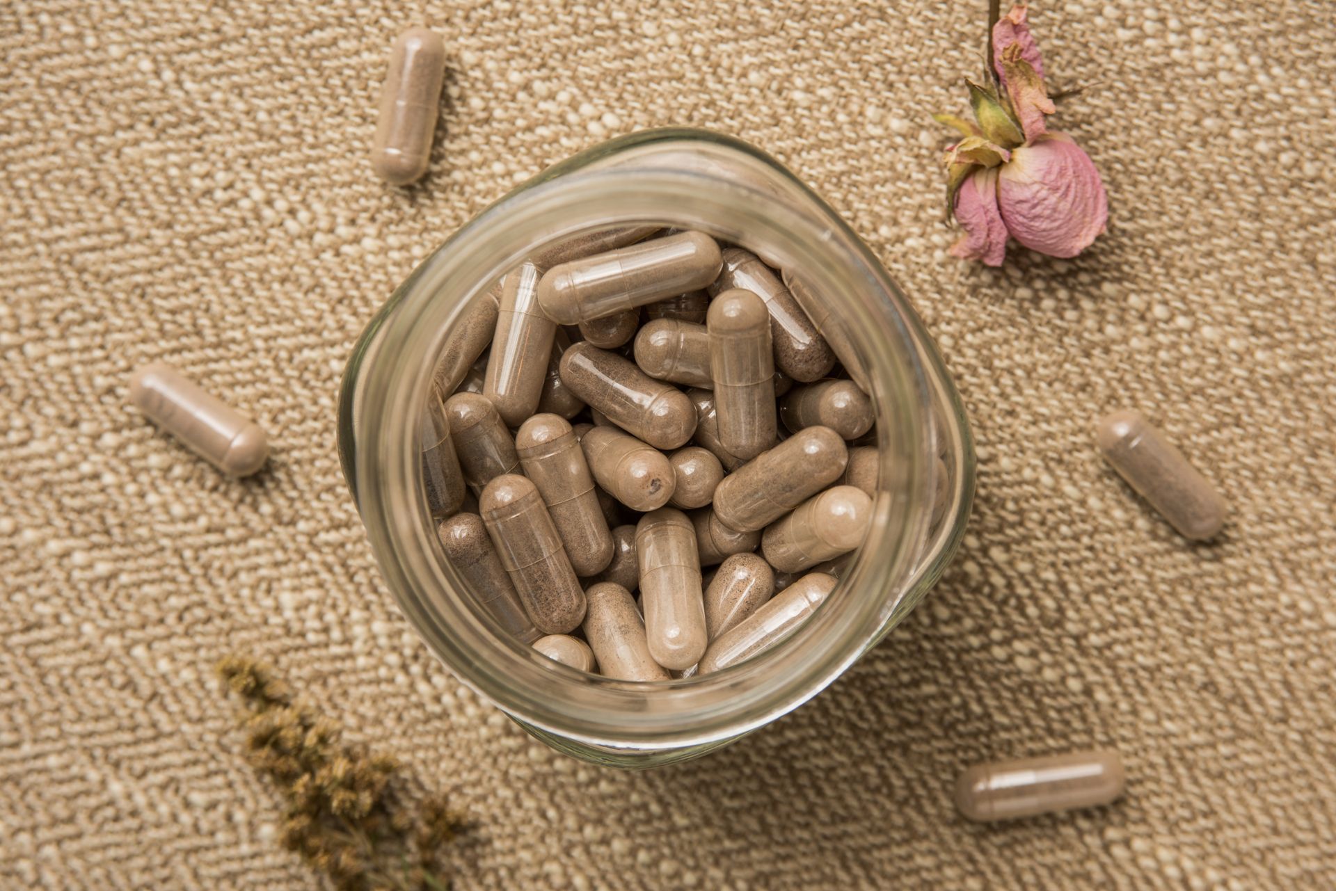 Brown capsules in a clear jar, scattered on a textured, beige surface with dried flowers.