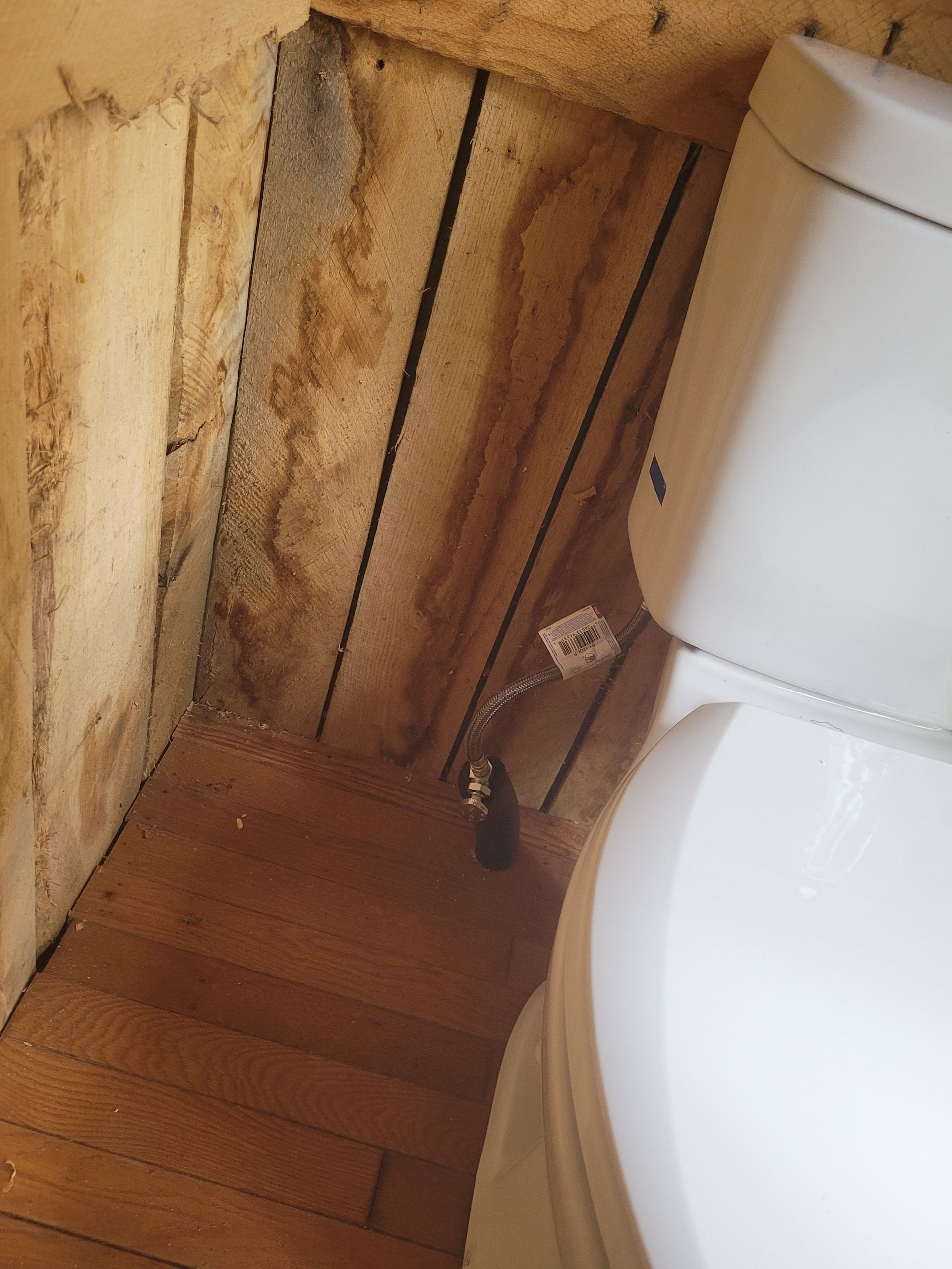 Toilet next to stained wooden wall and floor; water supply line visible.
