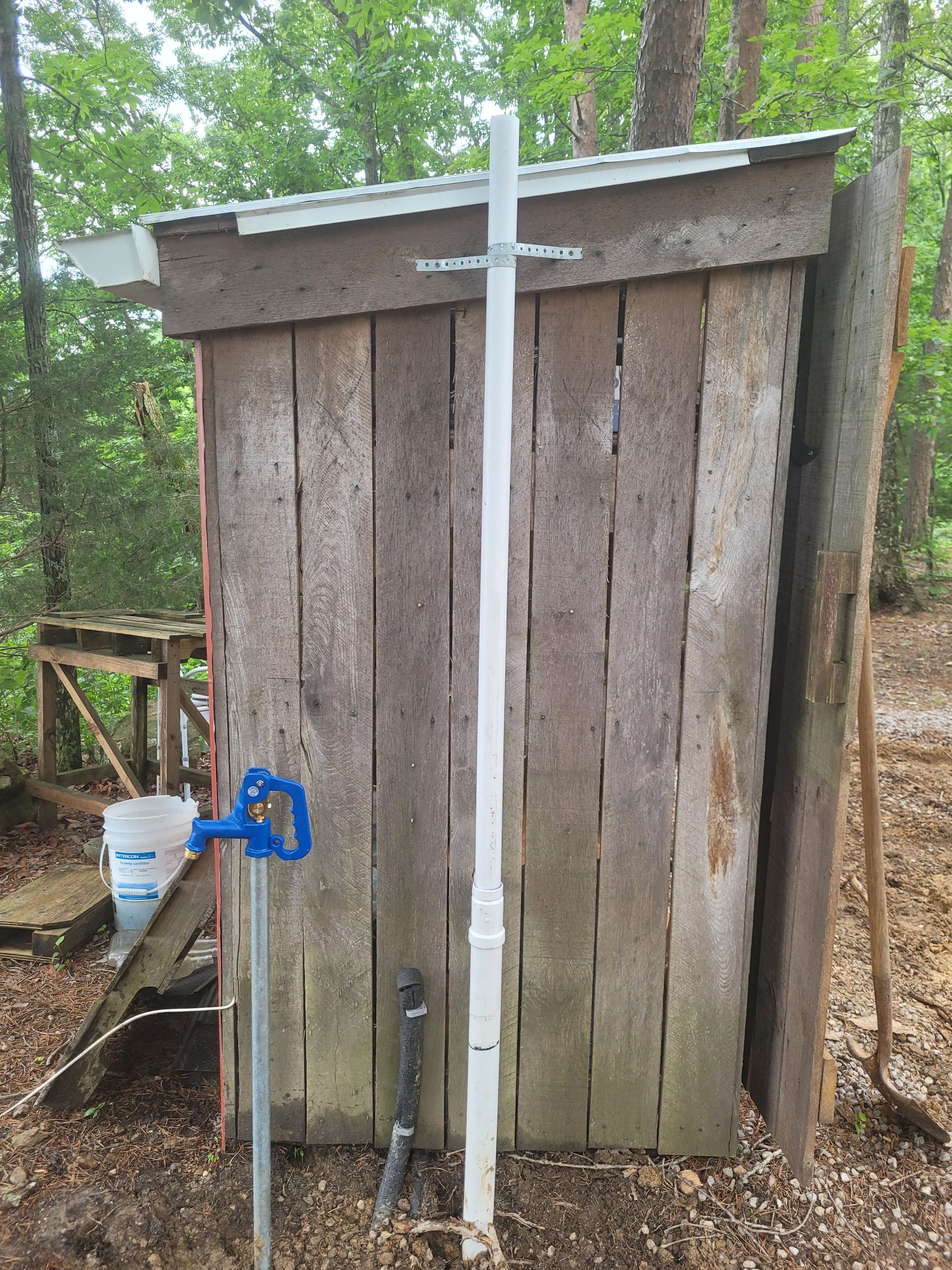 Wooden outhouse with a tall white pipe, blue water spigot, and bucket outdoors.