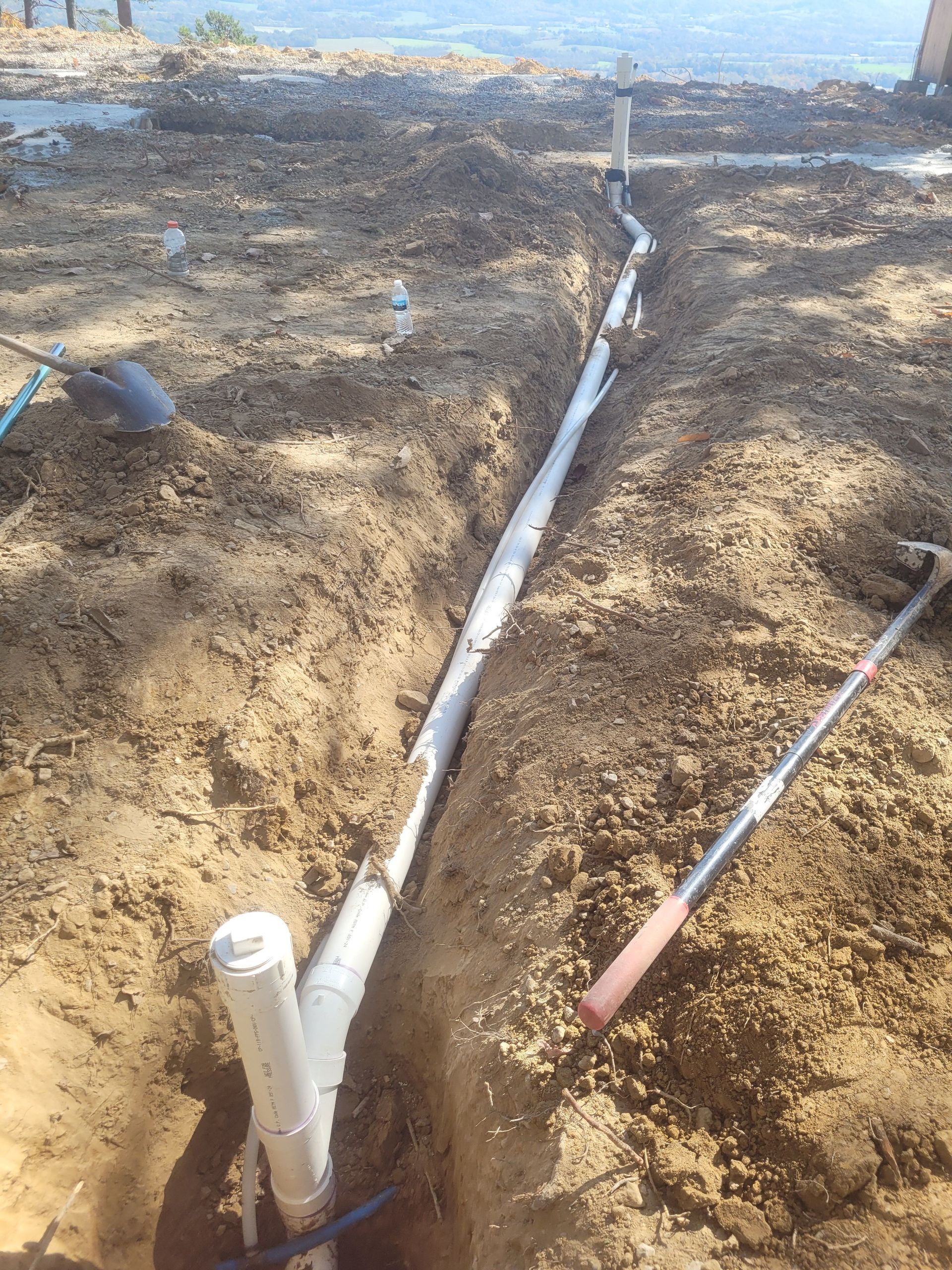Trench with white PVC pipes laid in the dirt, plumbing work in progress; a shovel and hammer lie nearby.