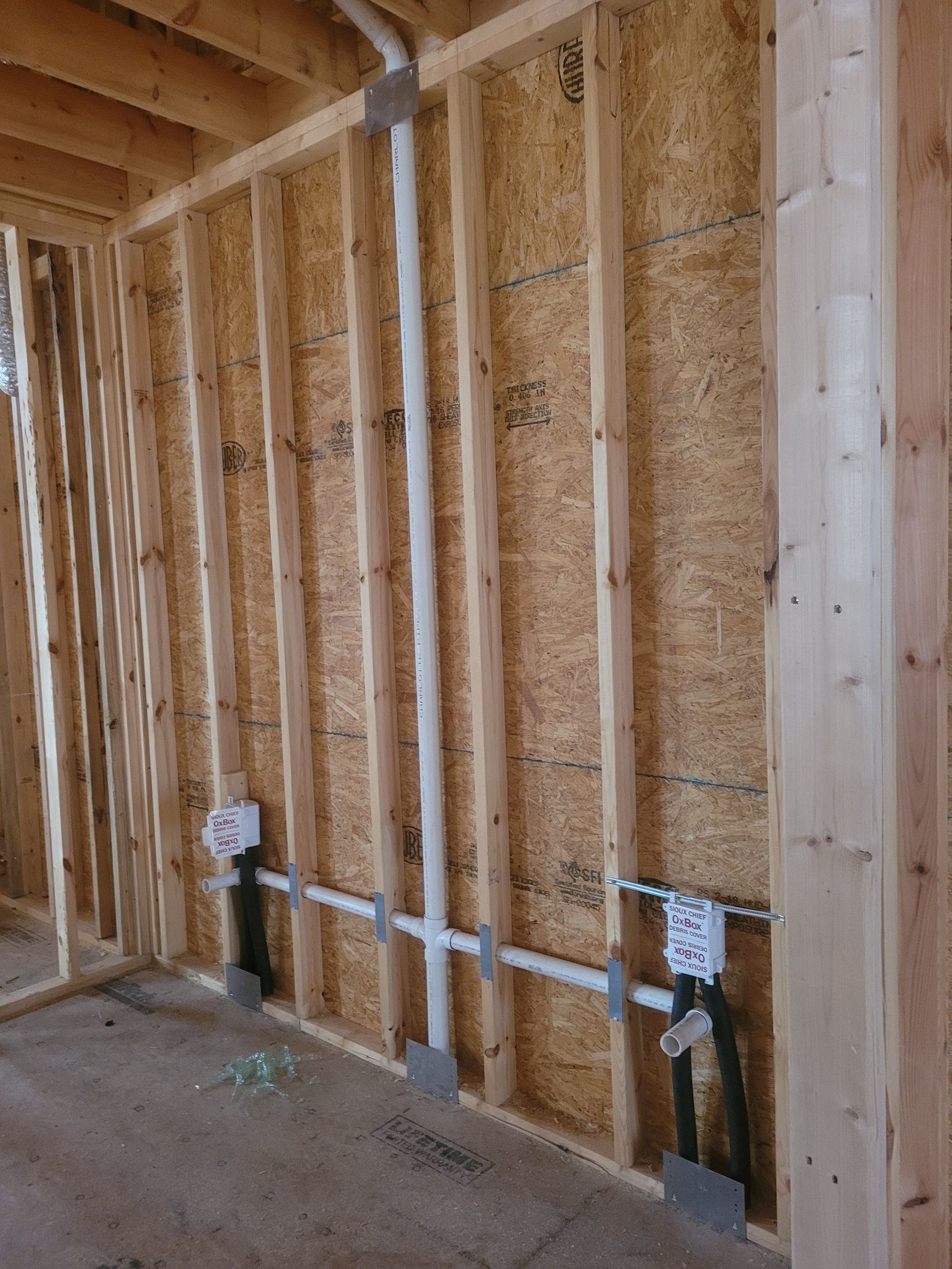 Interior wall of a building under construction, featuring wooden studs, plumbing pipes, and electrical boxes.