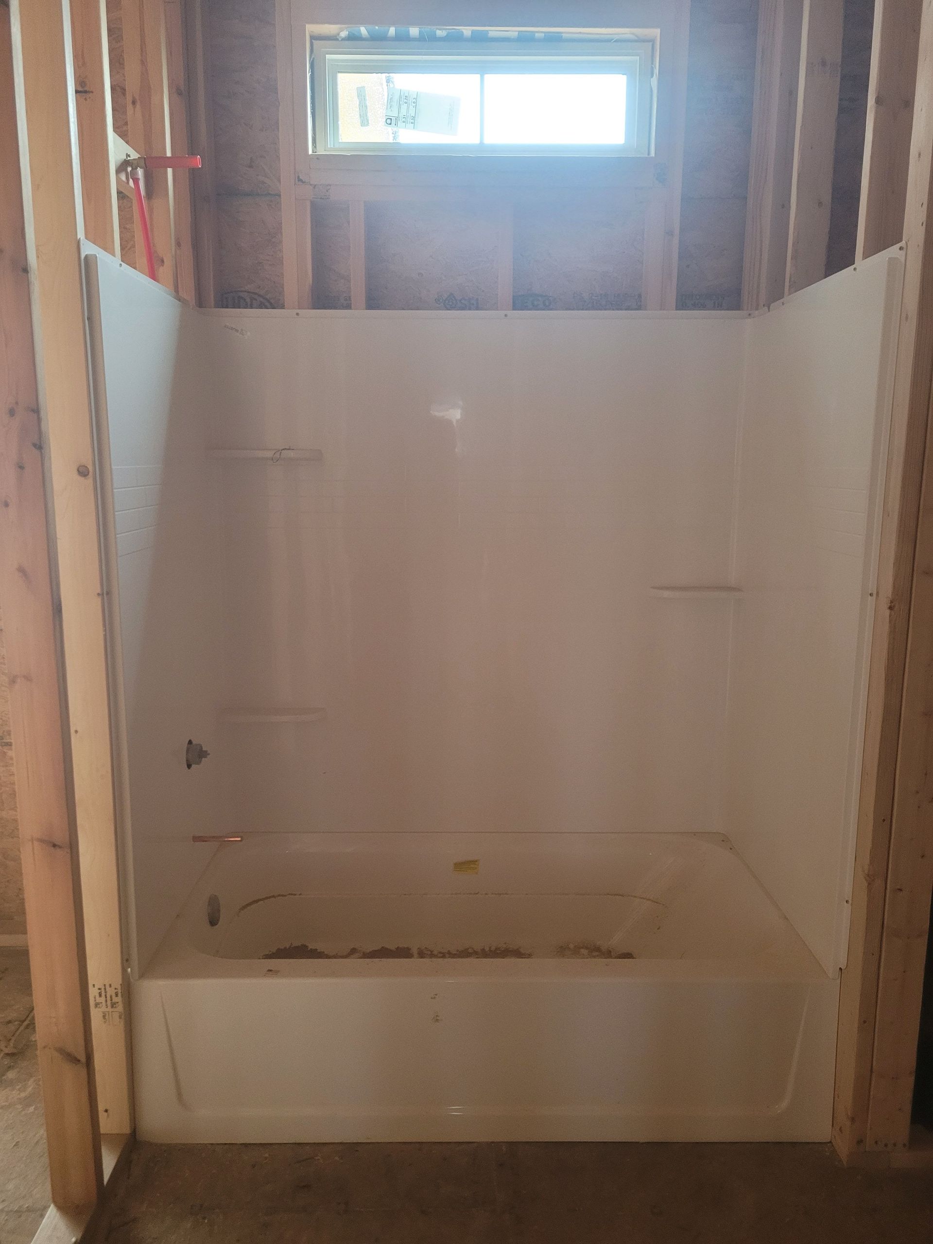 Interior shot of a white bathtub/shower unit framed in wooden studs, beneath a small window.
