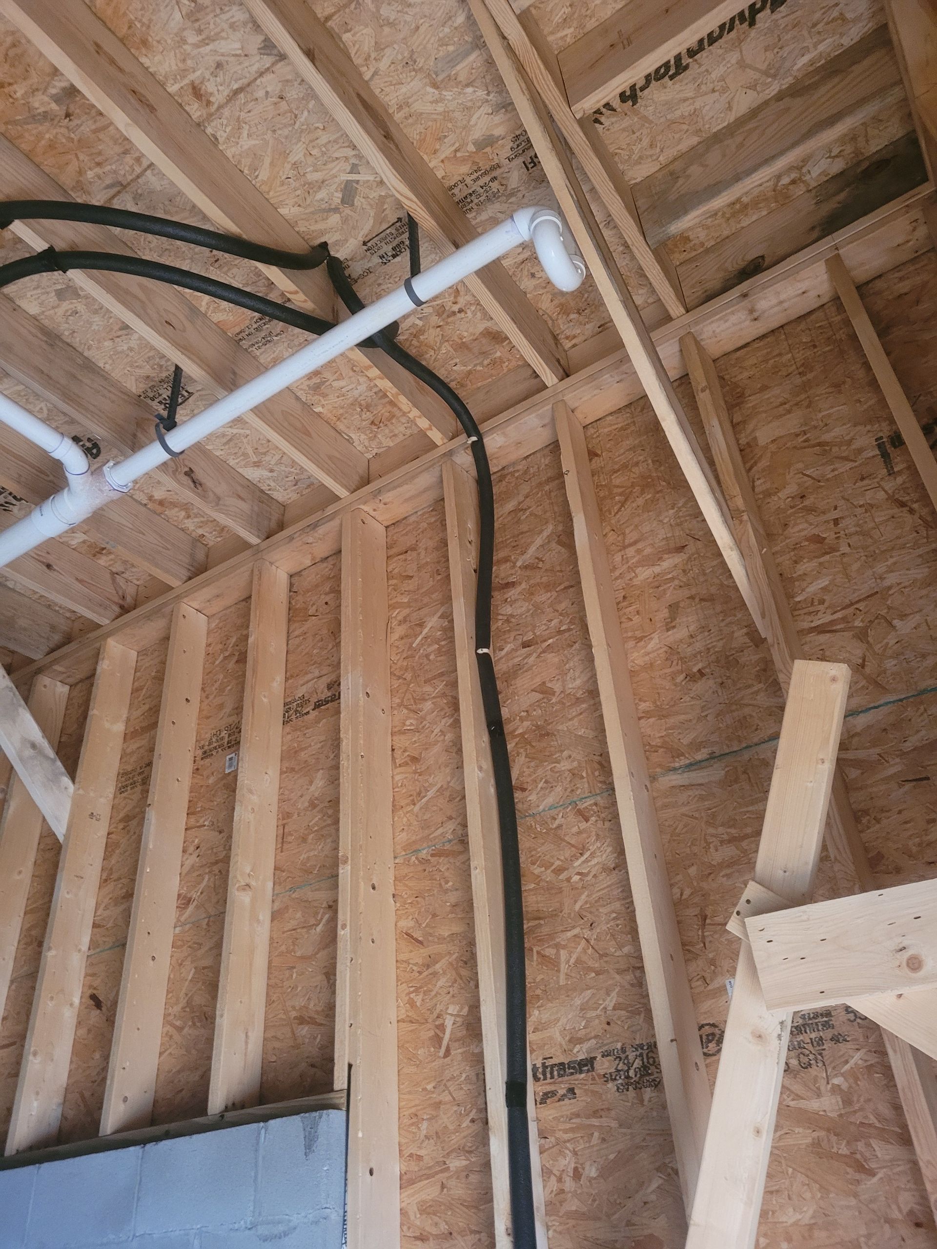 Interior view of a building under construction. Framing, black tubing, and white pipes are visible in a wooden structure.