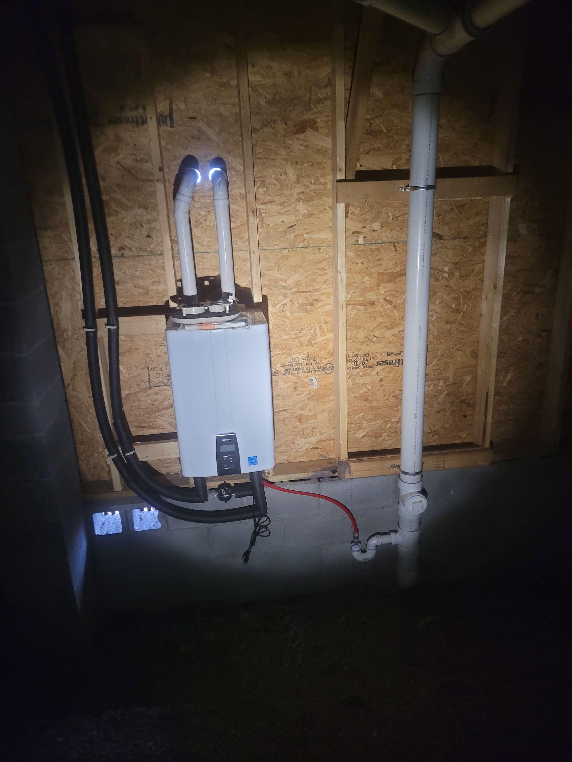 A wall-mounted, white tankless water heater with two white vent pipes, black hoses, and a red water line against wooden wall.