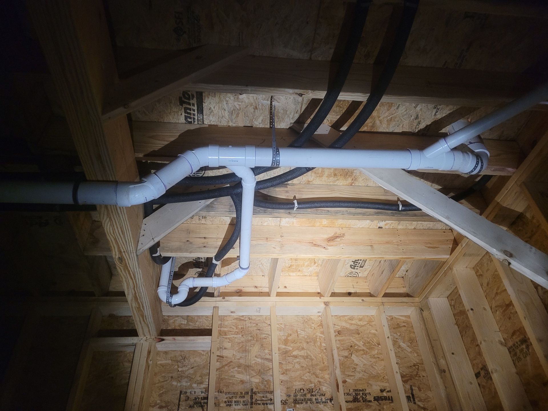 Plumbing pipes in a wood-framed ceiling. White and black pipes are connected, visible in a new construction setting.