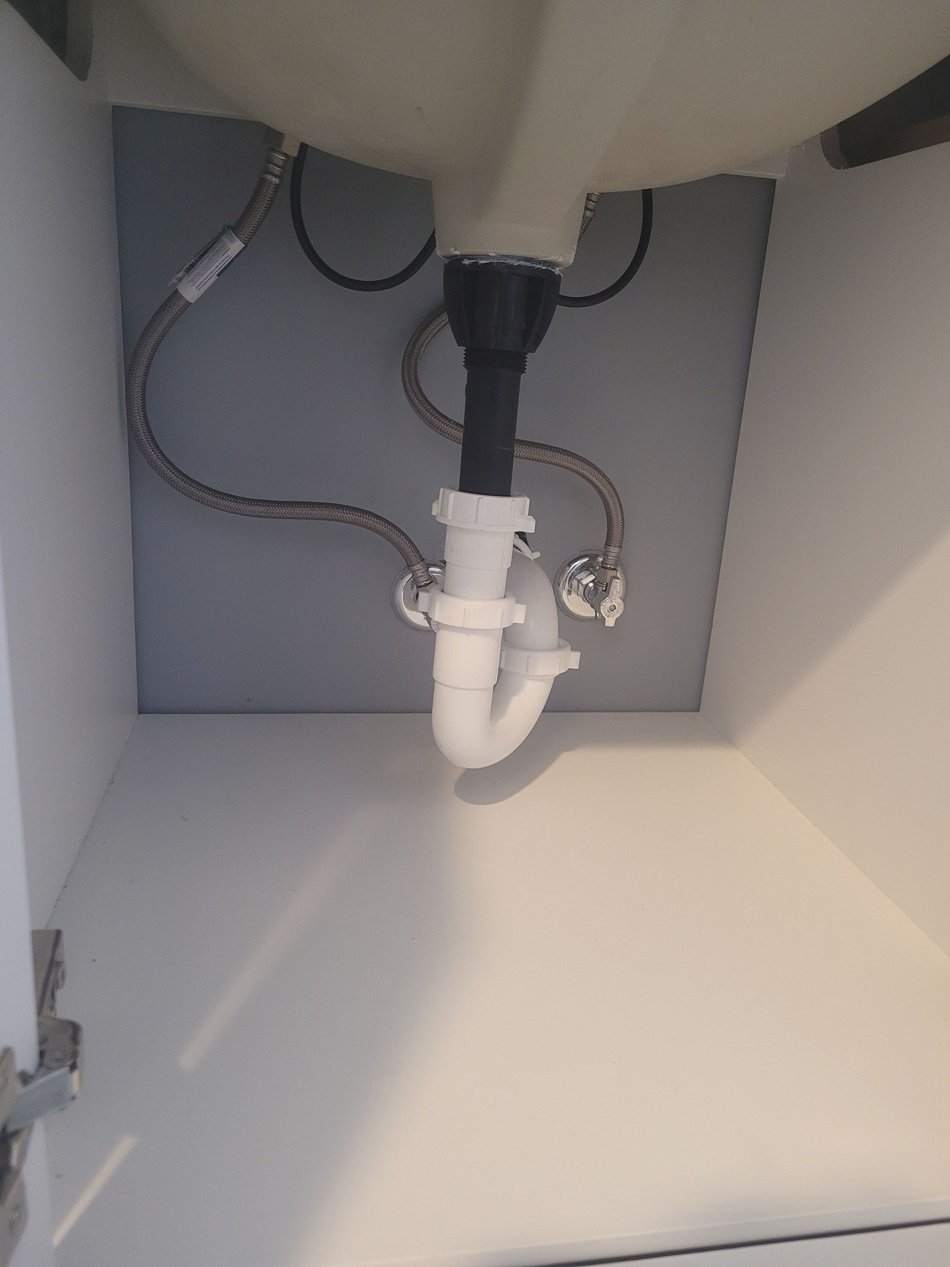 Under-sink plumbing: white pipes, curved trap, chrome supply lines, and a dark drainpipe against a gray cabinet interior.