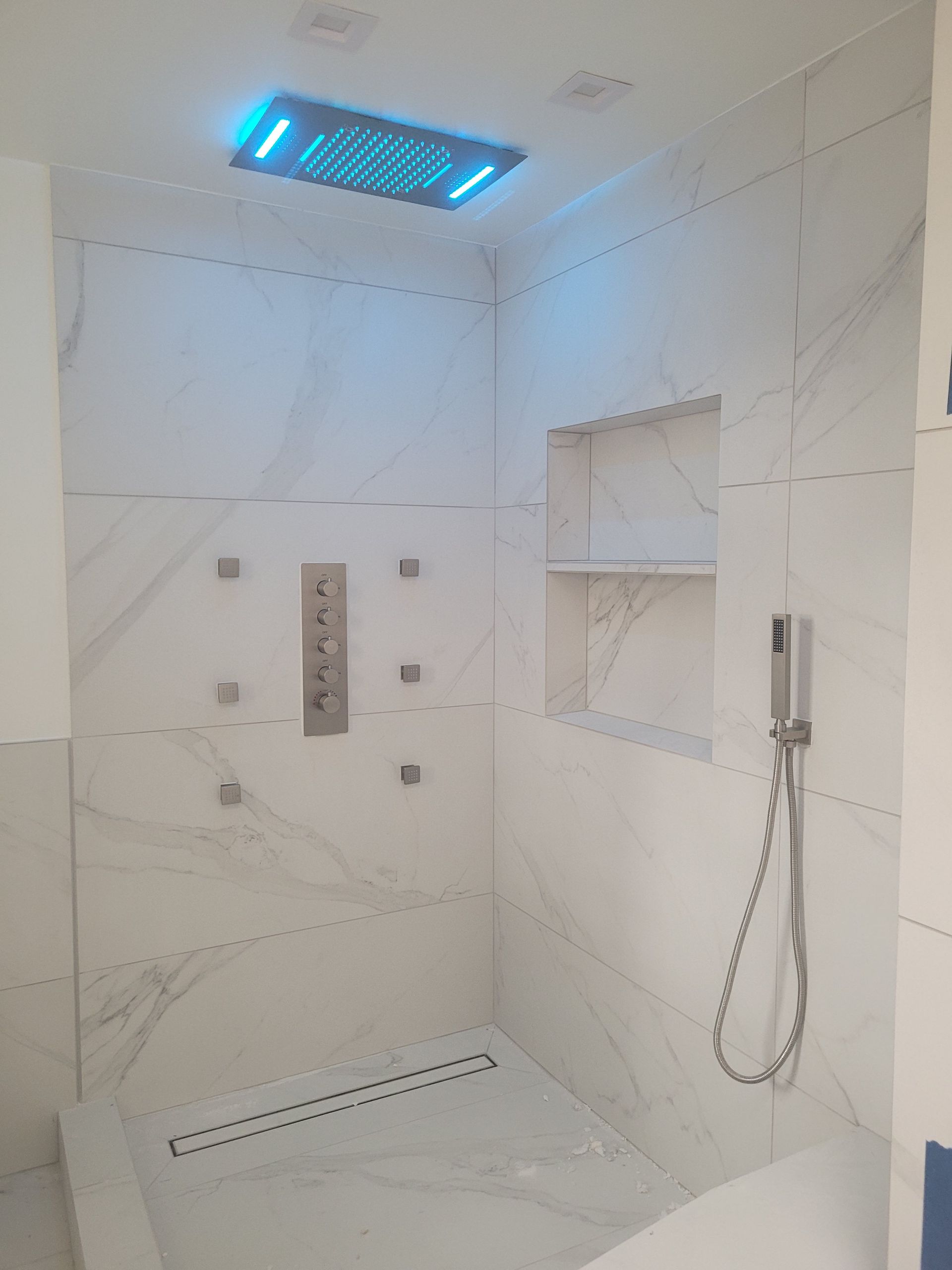 White-tiled shower with blue overhead light and built-in shelving and controls.