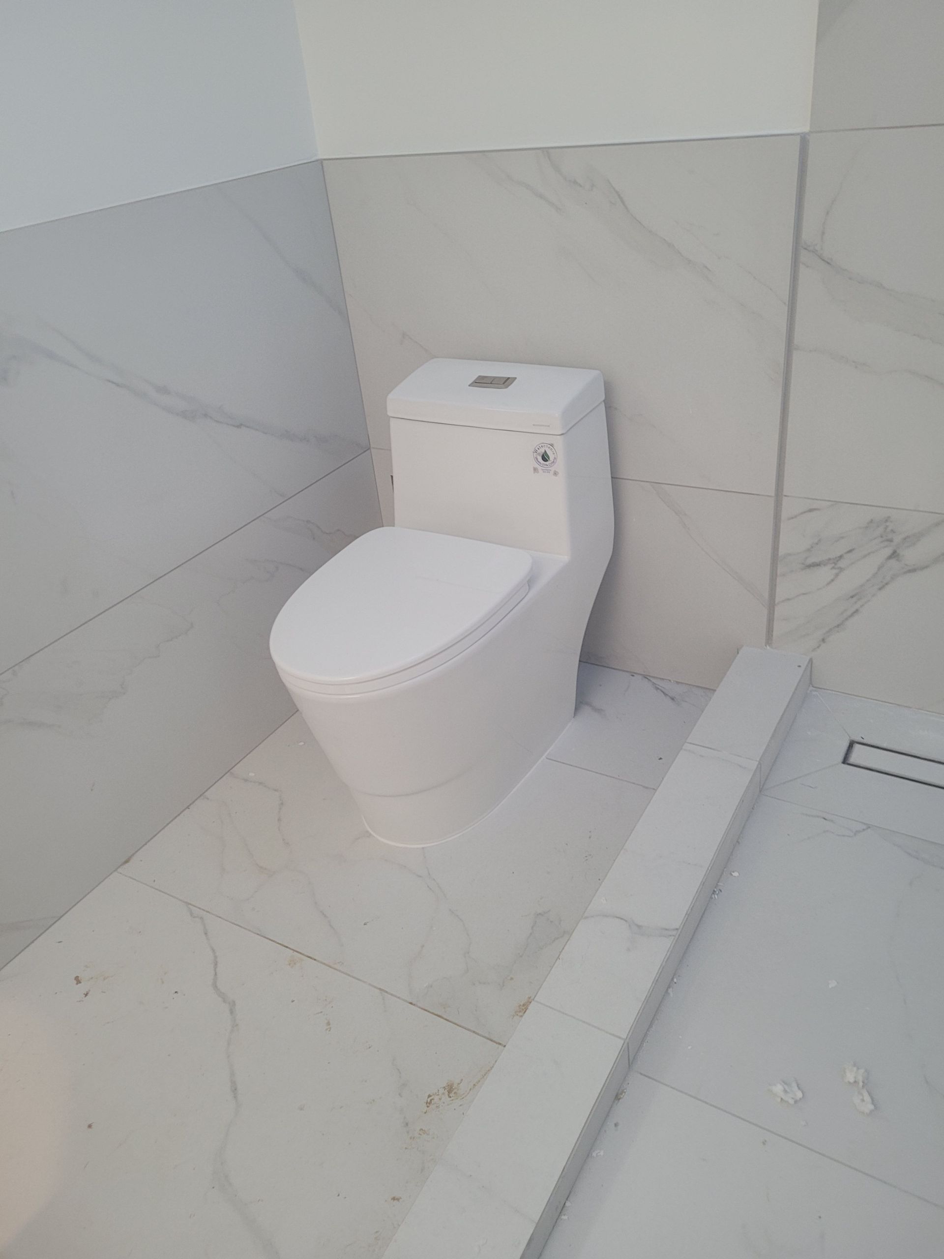White toilet in a bathroom with marble-patterned tile walls and floor; a low, gray structure is in the foreground.