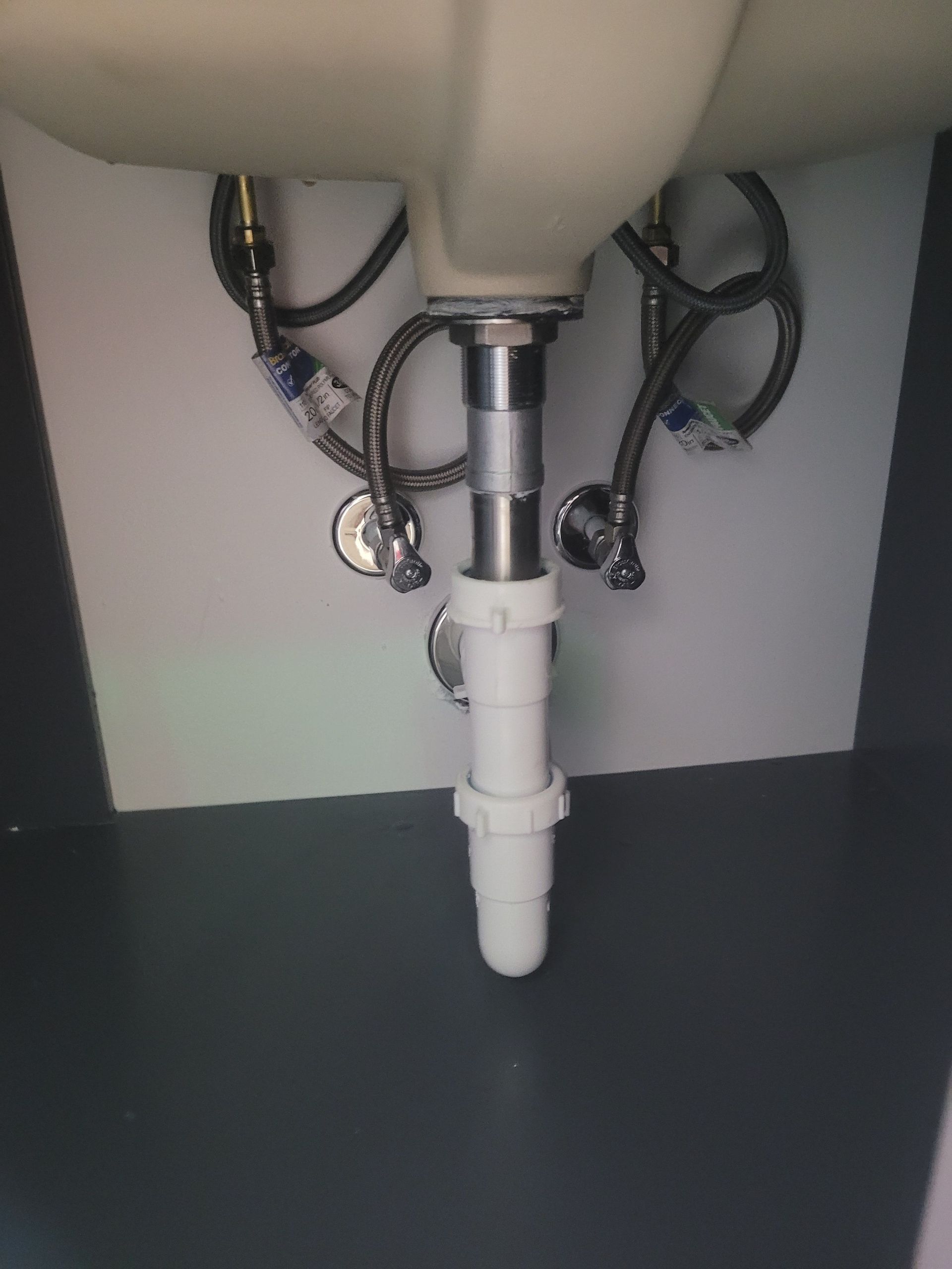 Underside view of a white bathroom sink with chrome drain pipes and flexible water supply lines. White cabinet is visible.