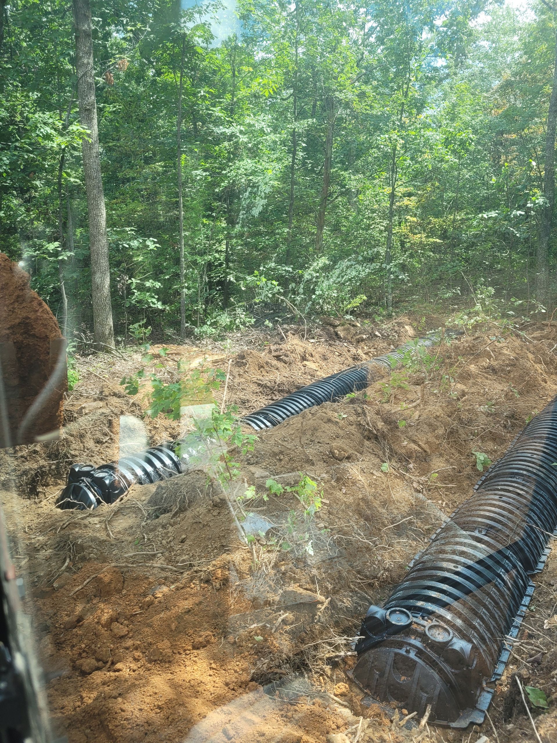 Two black drainage pipes in a dirt trench, surrounded by brown soil and green trees.