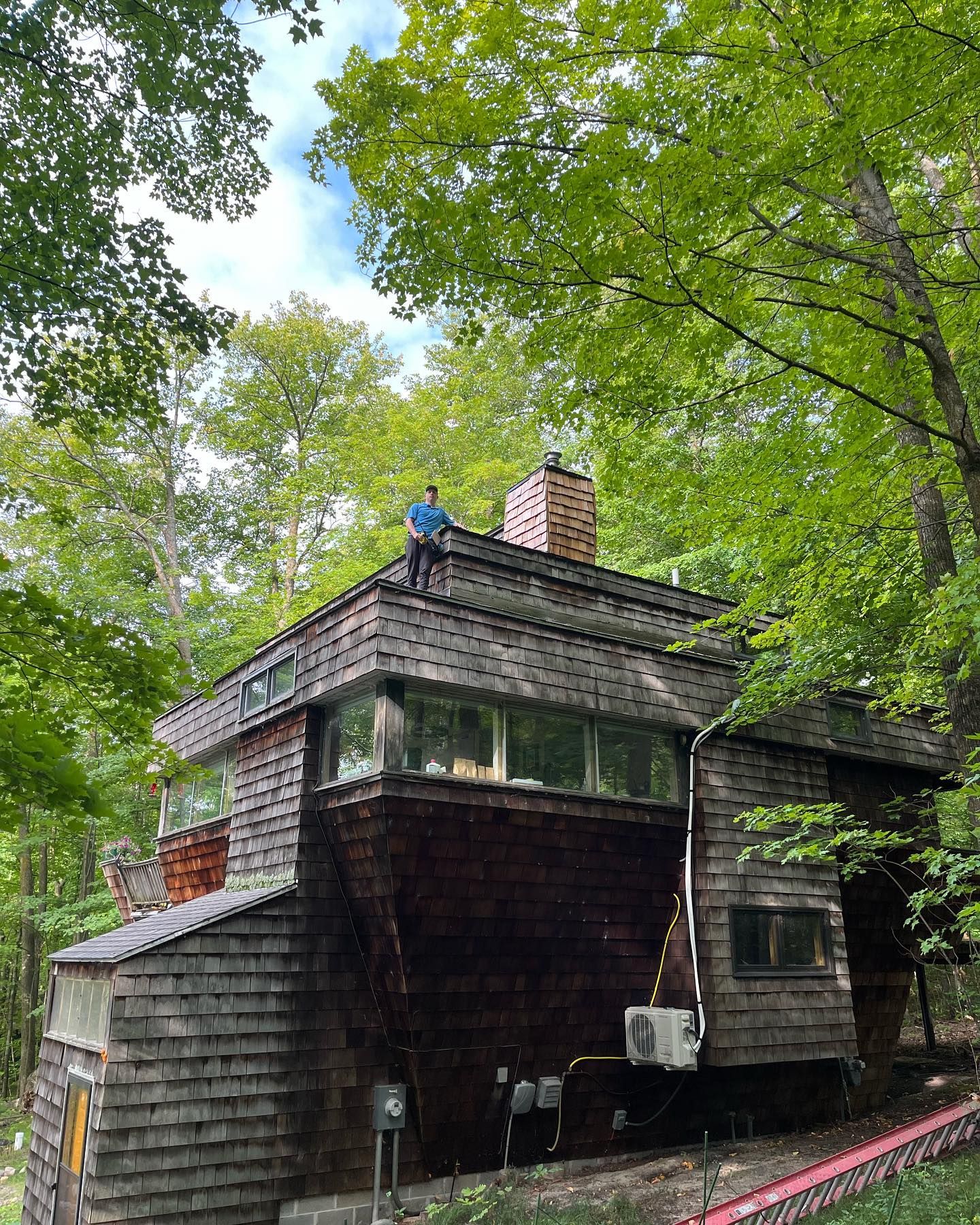 Person on the roof of a wooden house surrounded by trees on a bright day.