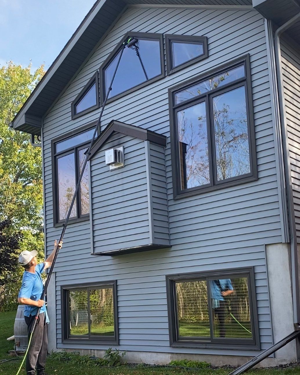 Person cleaning windows of a gray house with a water-fed pole. Sunny outdoor setting.