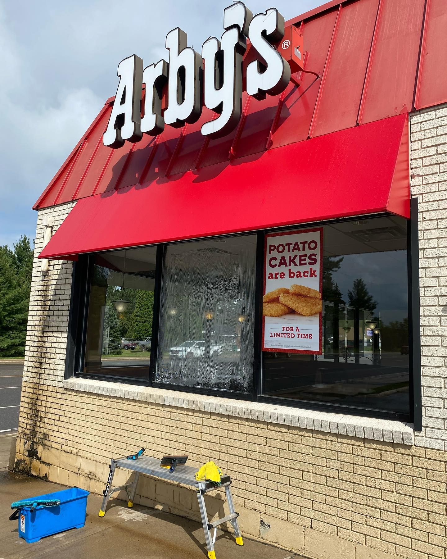 Arby's restaurant exterior with red awning, brick facade, and sign advertising potato cakes. Cleaning equipment visible below windows.