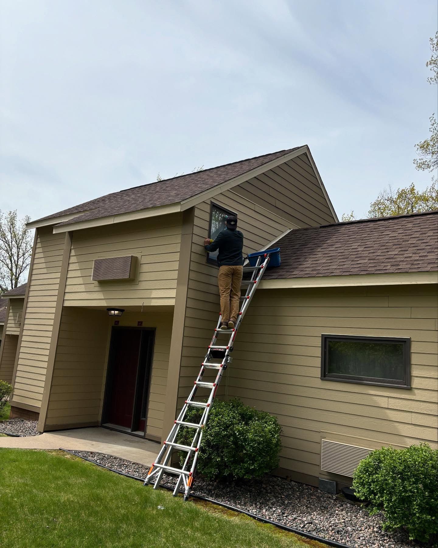 Person on ladder working on the exterior of a tan building with a brown roof.