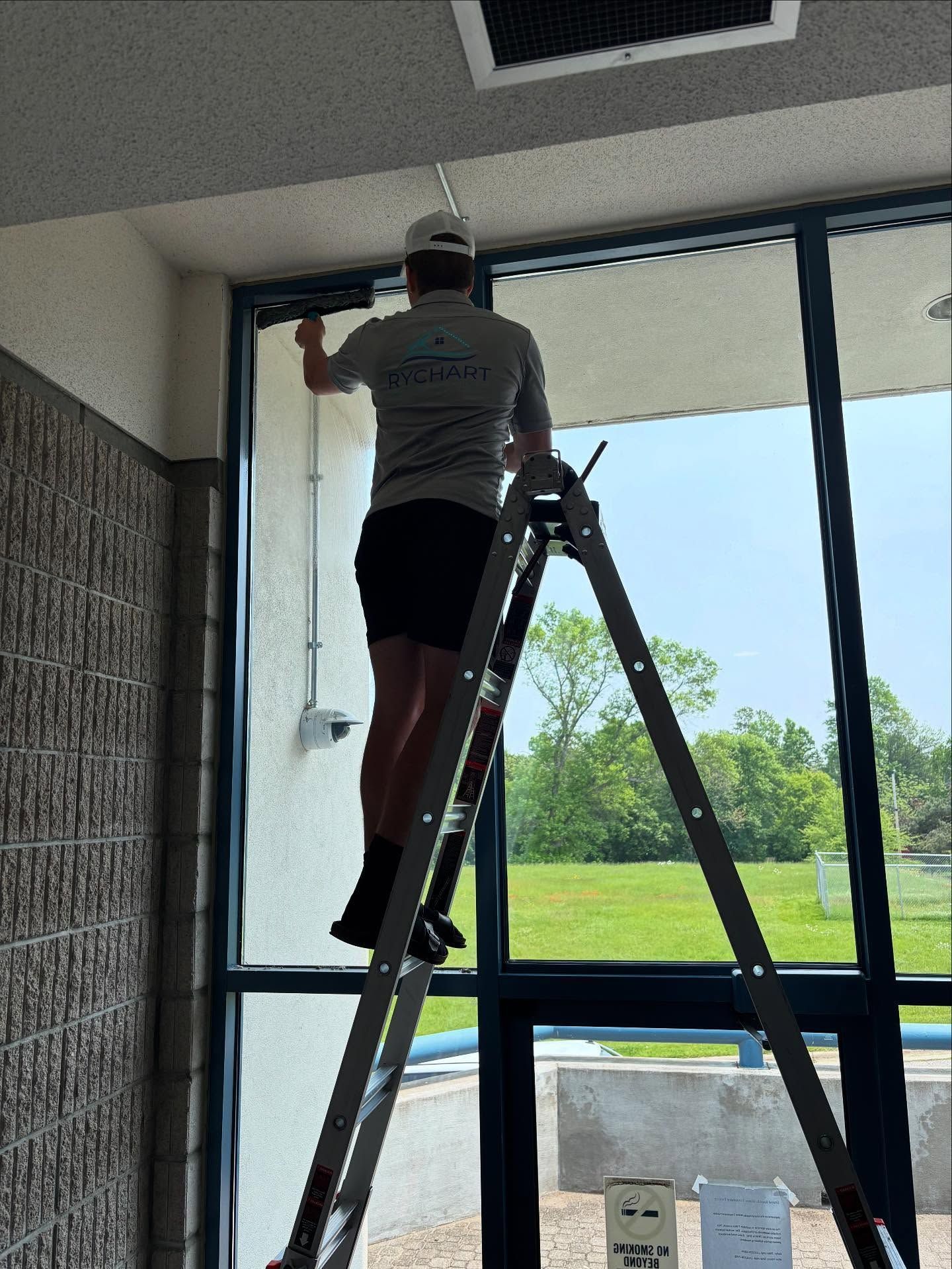 Man on ladder cleaning a large window. He's wearing shorts and a shirt, with a view of green grass and sky outside.