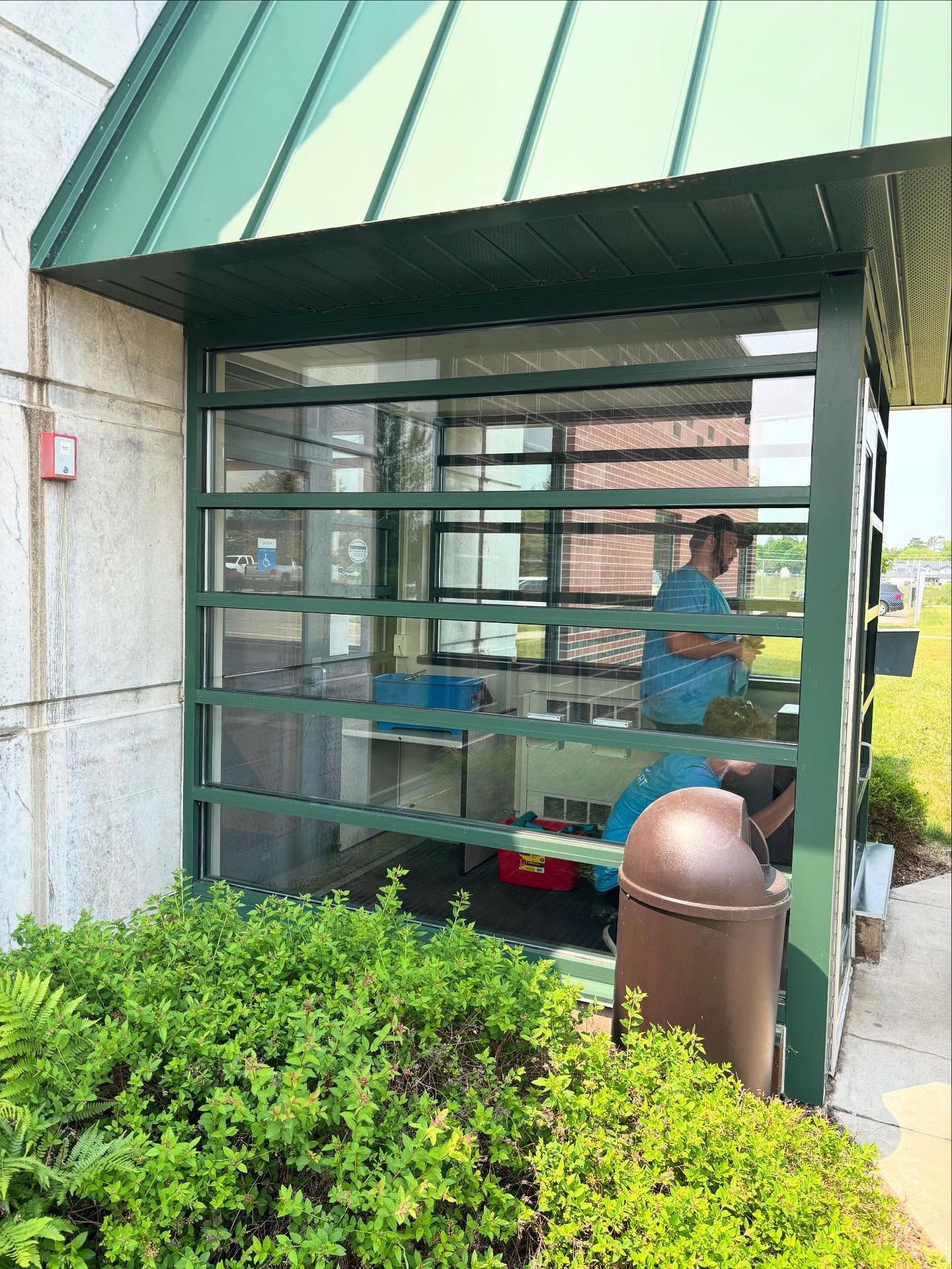 Green-framed glass window with a person inside, under a green roof. Bushes and a trash can in front.