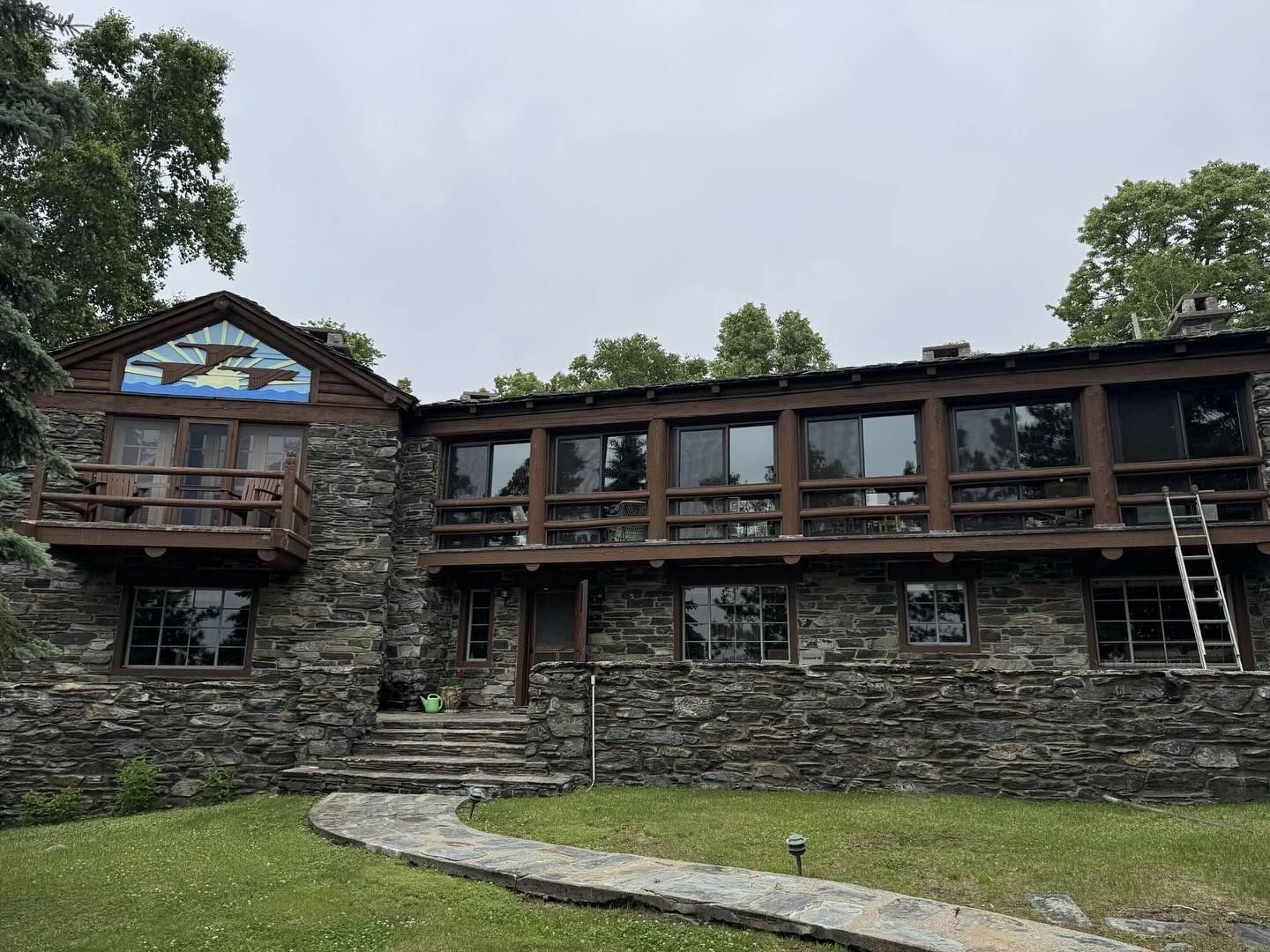 Stone building with a wooden balcony, steps, and a ladder. A blue-toned mural is above a doorway. Overcast sky.