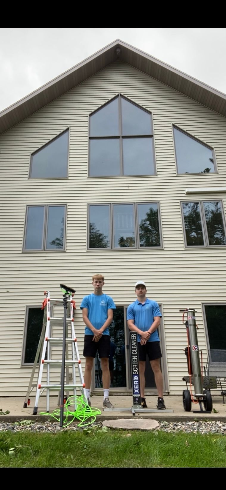 Two men in blue shirts and shorts stand in front of a house with ladder and equipment.