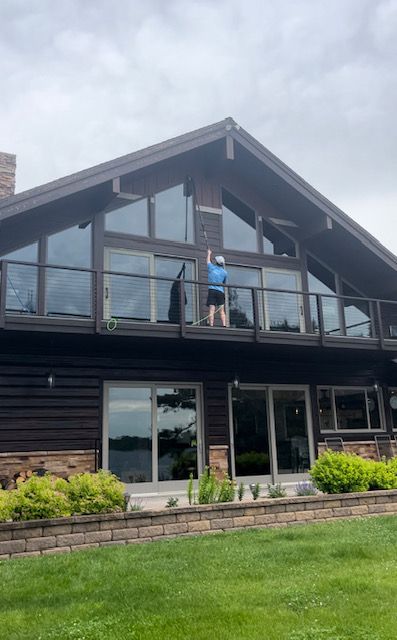 Person cleaning windows of a large house with a balcony, standing outside under a cloudy sky.