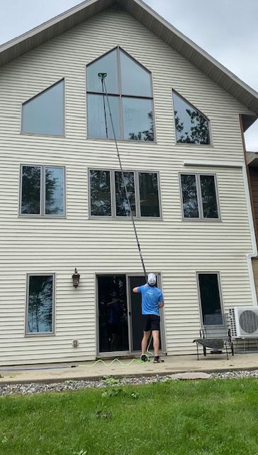 Man cleaning windows on a two-story beige house with a long pole. Overcast day, green grass.