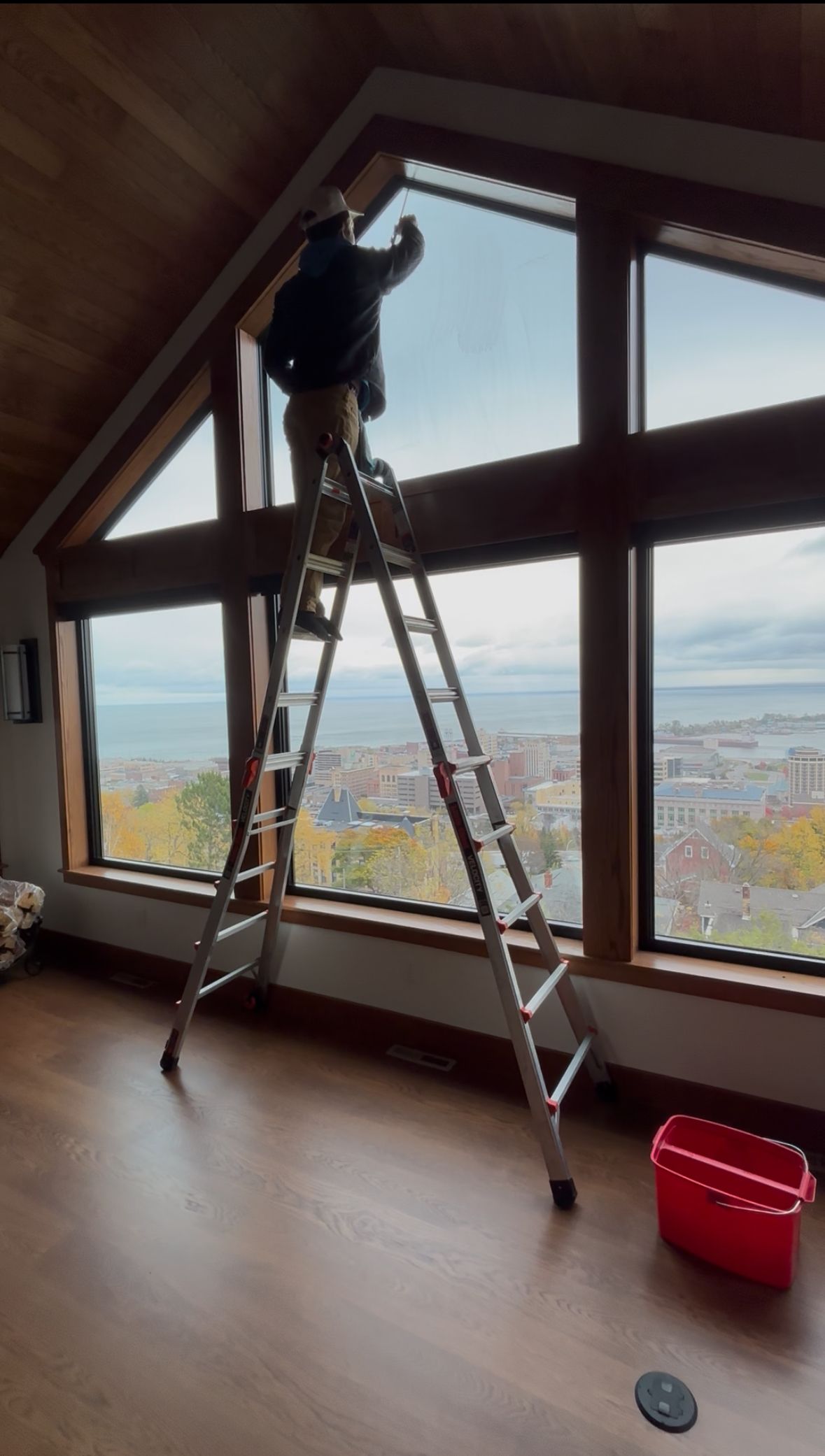 Person cleaning windows of a gray house with a water-fed pole. Sunny outdoor setting.