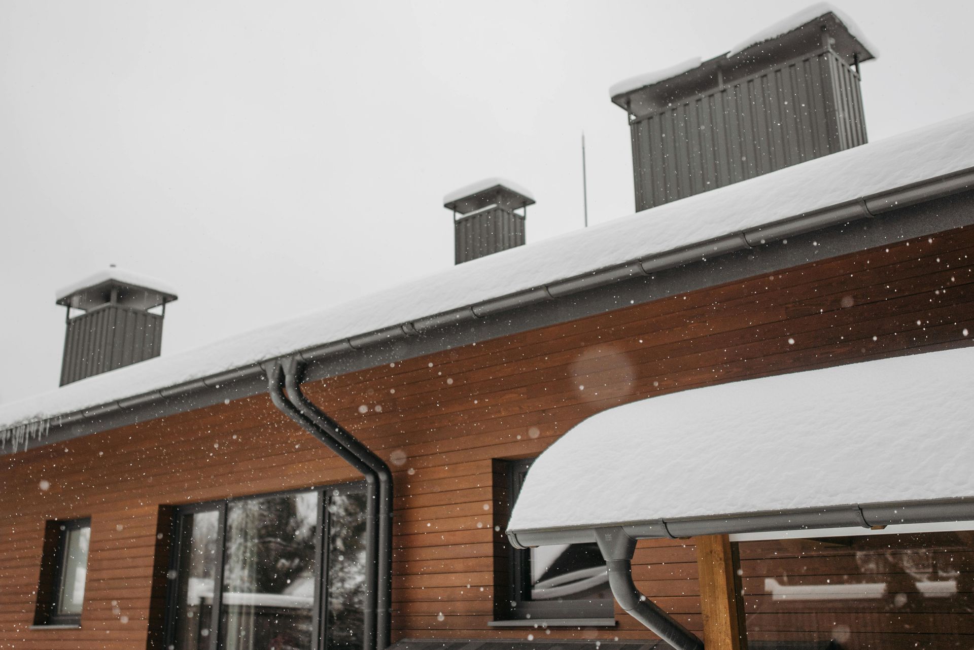 Snowy roof of a wooden building with three chimneys and a gutter, falling snow.
