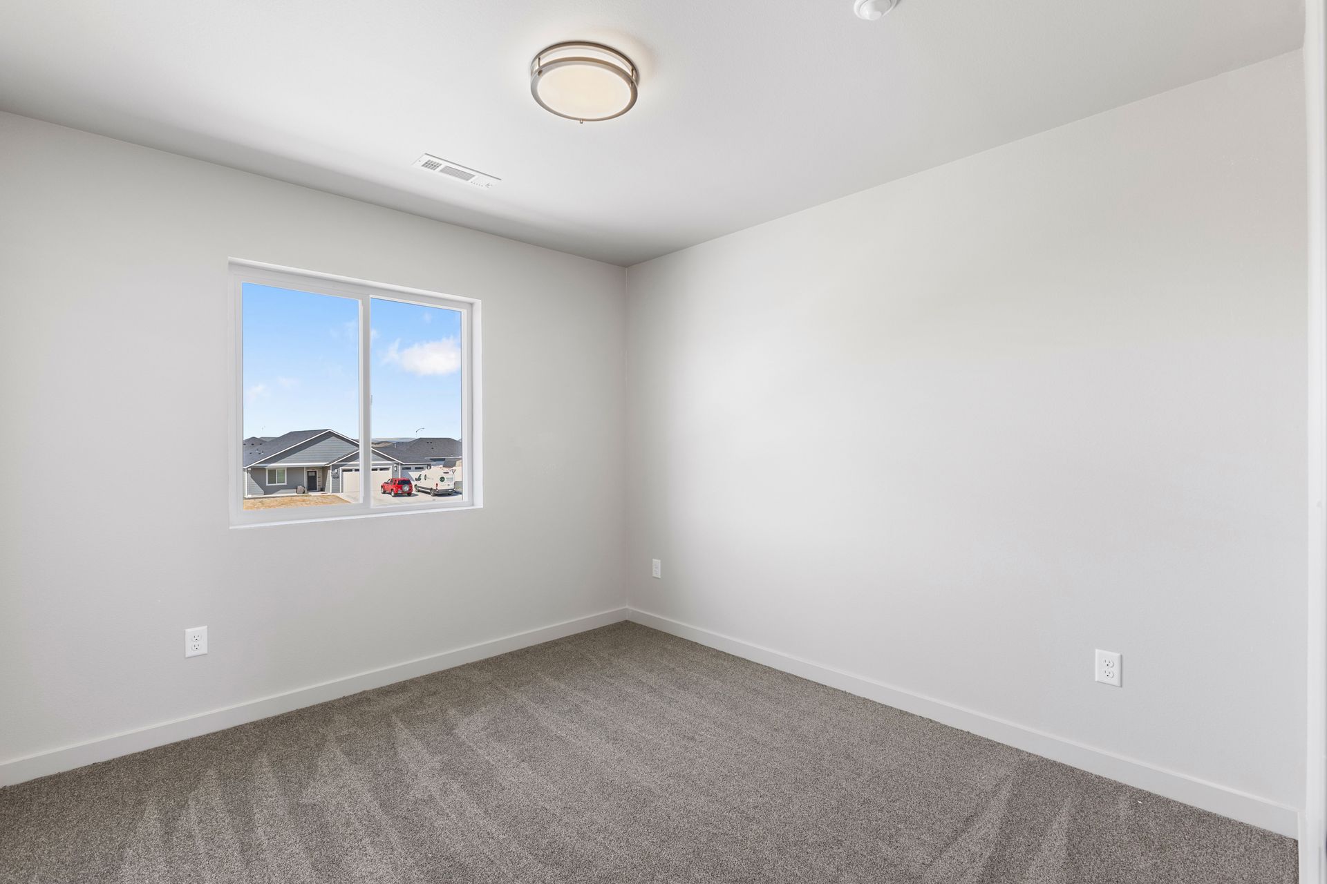 Empty bedroom with gray carpet, white walls, and a window overlooking houses.