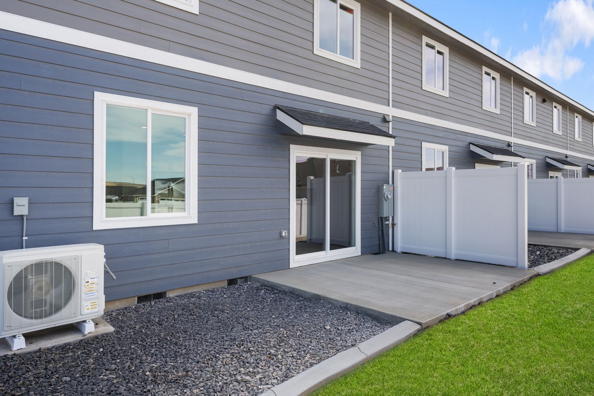 Exterior of a gray building with white trim, patio, and air conditioner. Green grass and dark gravel.