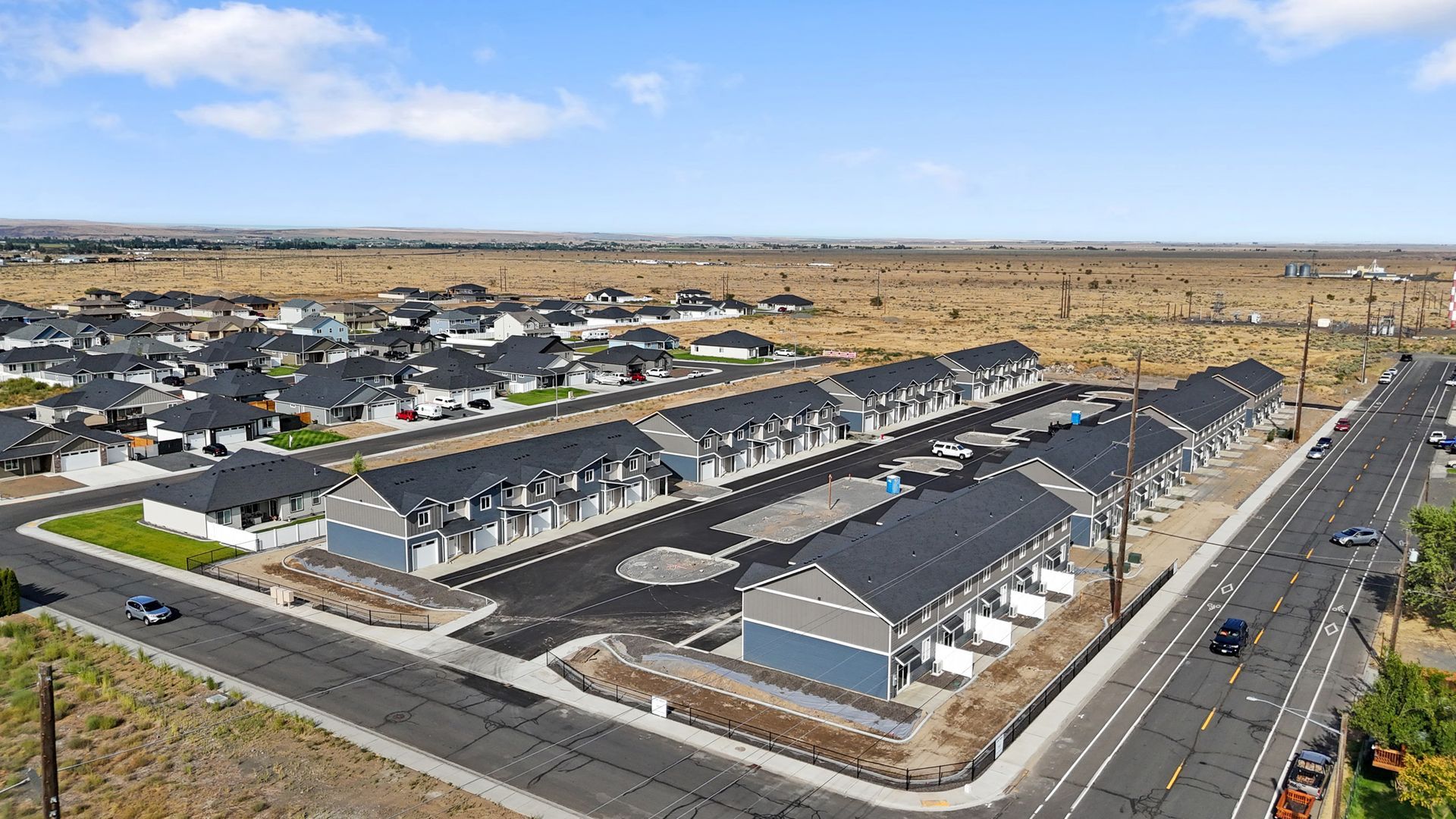 Aerial view: rows of townhomes, gray roofs, light blue siding, black asphalt, street, rural setting.