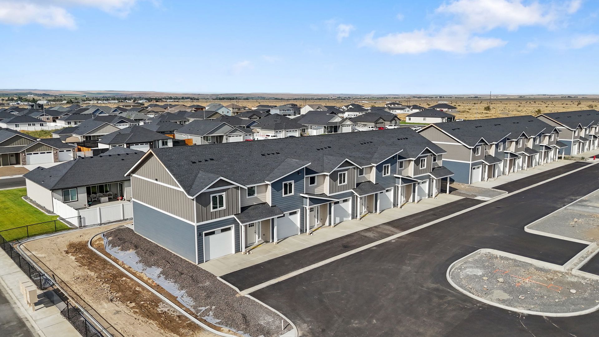 Row of gray townhomes with black roofs and attached garages, in a new residential neighborhood.