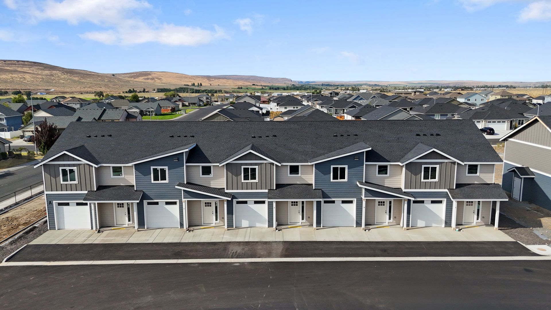 Row of new townhouses, with gray and blue siding, dark roofs, and white garage doors, in a suburban setting.