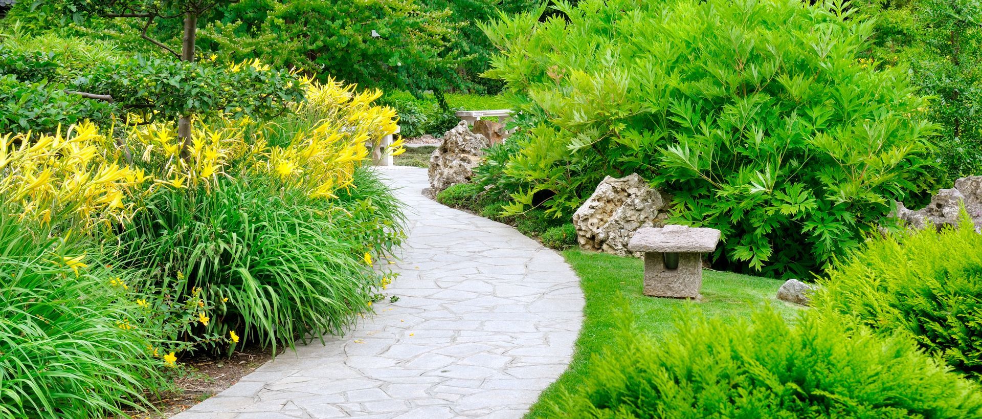 A winding stone path through a lush green garden, with yellow flowers and a small decorative birdhouse.