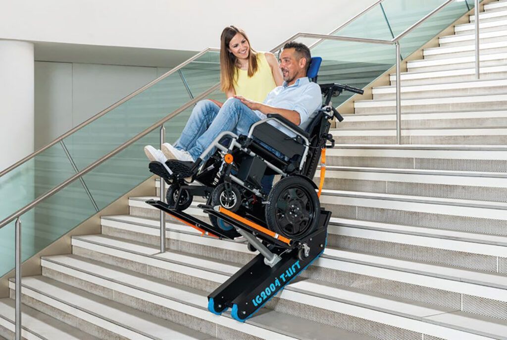 Un utente su sedia a rotelle viene aiutato a salire le scale tramite un dispositivo elettrico, mentre sorride all'assistente su una scala interna.