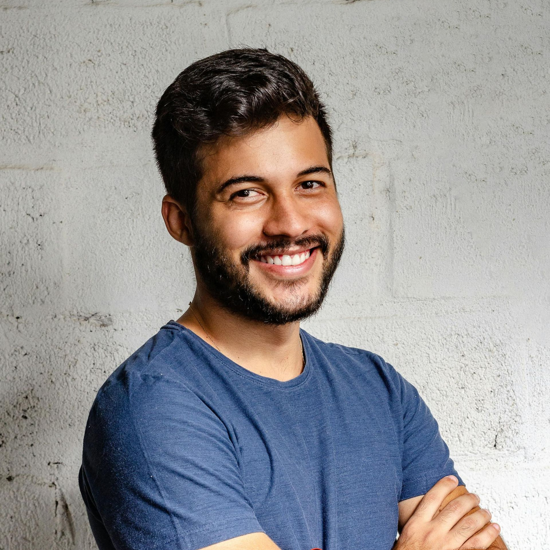 Un uomo con i capelli scuri e la barba sorride, con le braccia incrociate e una maglietta blu, contro un muro bianco ruvido.