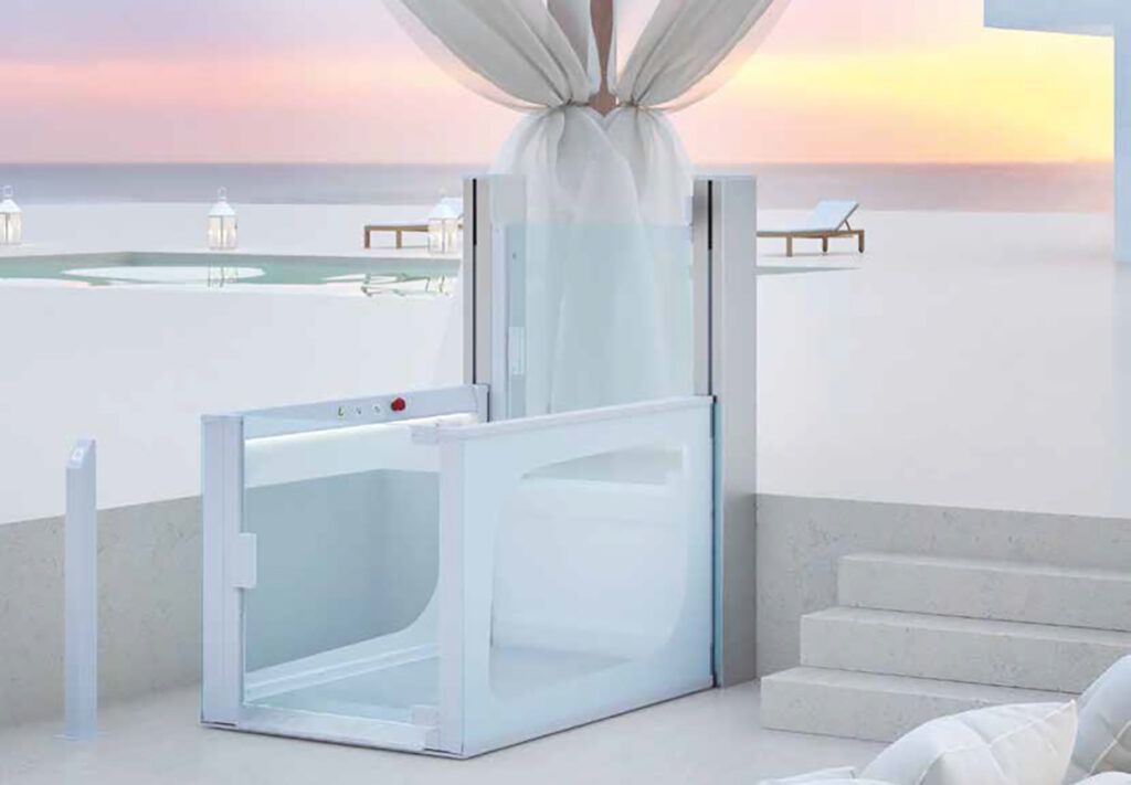 Ascensore esterno bianco con pareti in vetro su una terrazza con vista sulla spiaggia e sul mare.