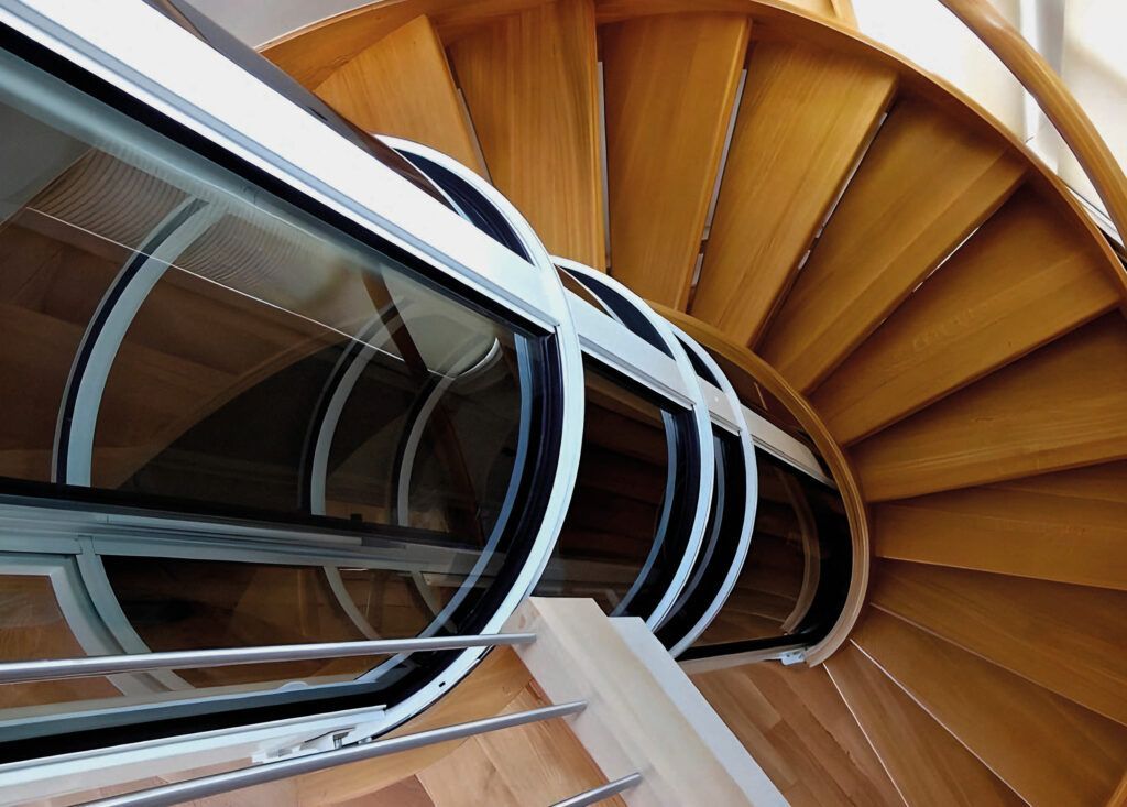 Scala a chiocciola con vano ascensore in vetro. Gradini e ringhiera in legno.