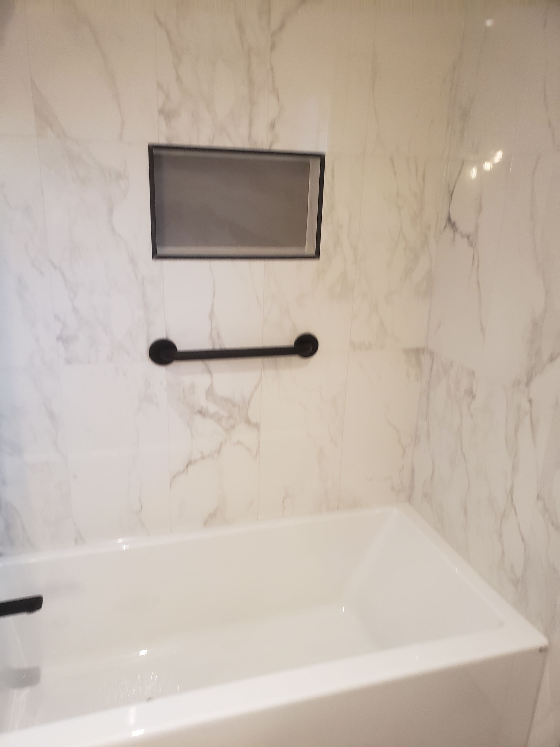 White tiled bathroom with black handrail and recessed shelf above a bathtub.