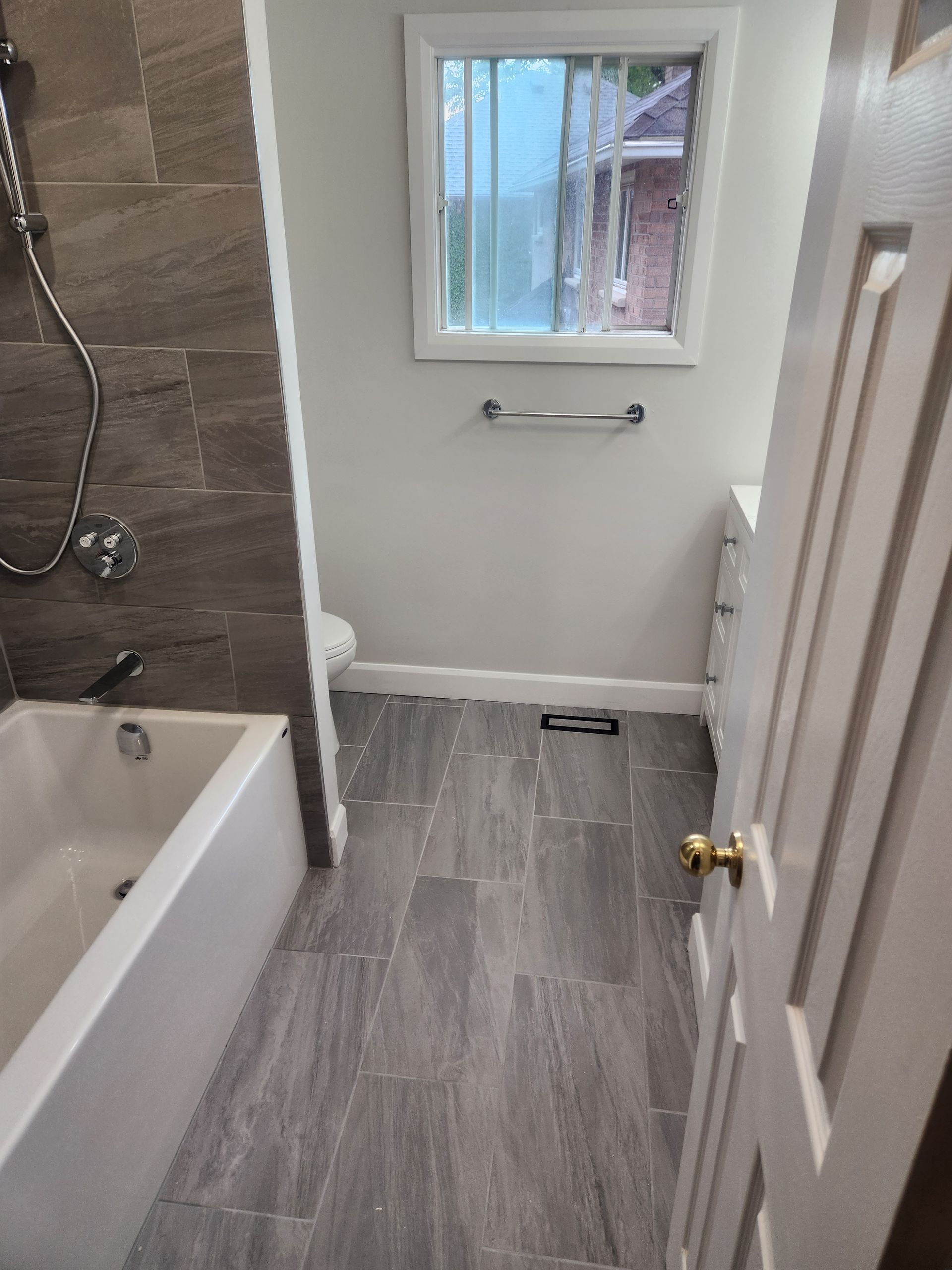Bathroom with gray tile floor, white tub and vanity, window. Gray tile wall behind shower.