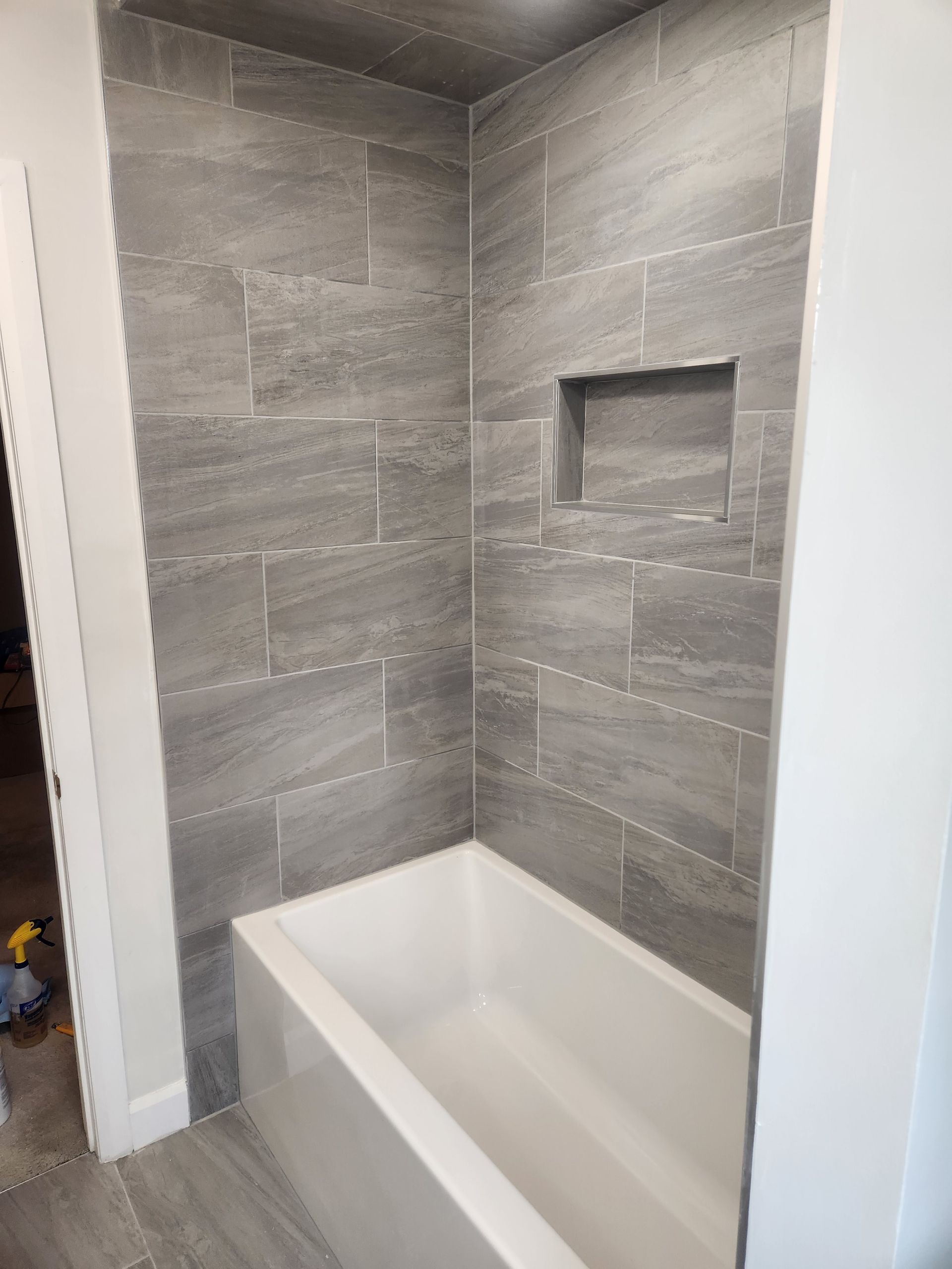 Gray tiled shower with white tub and built-in shelf.