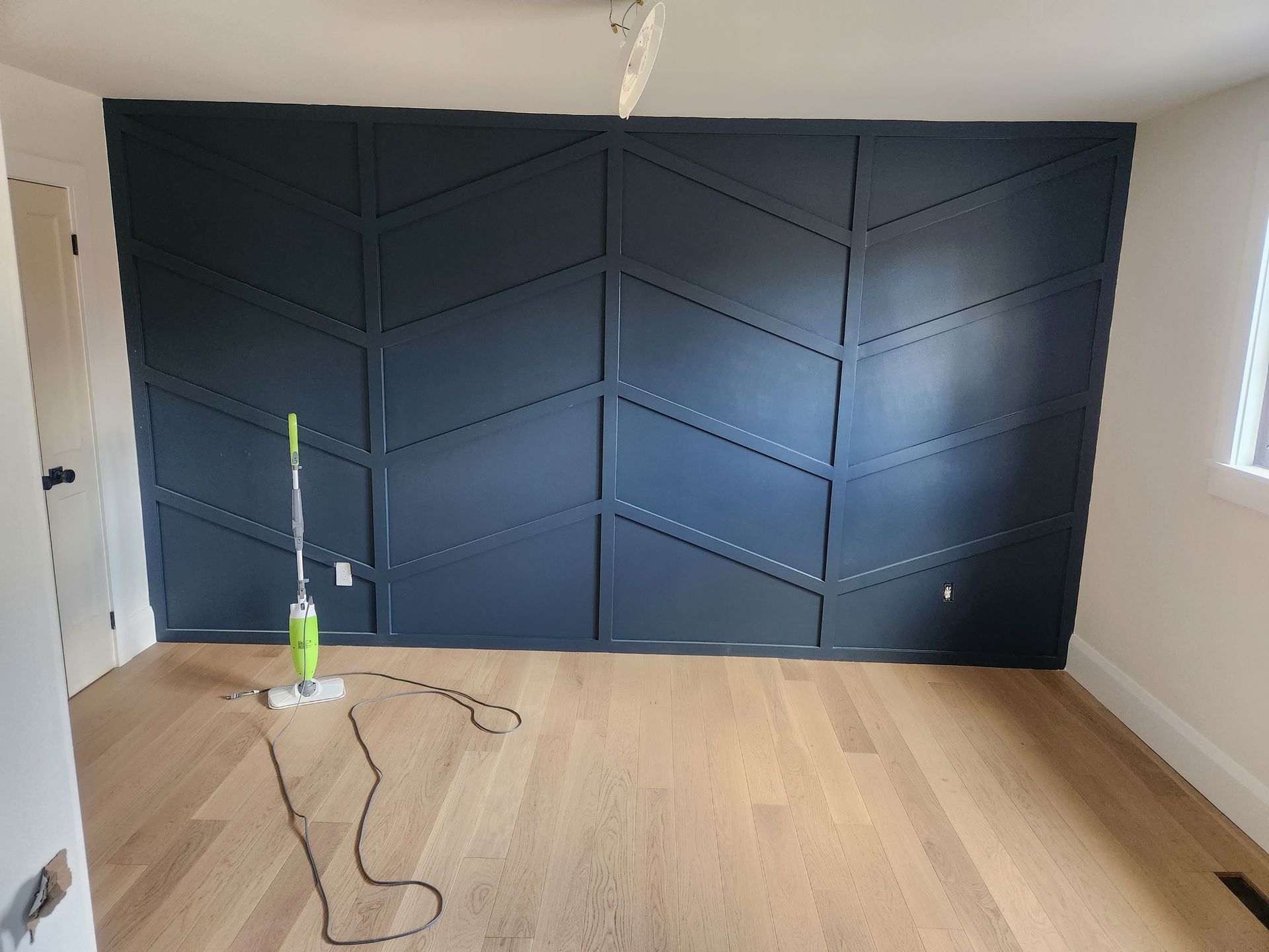 A navy blue accent wall with a geometric design in a room with wooden floors; a mop sits on the floor.