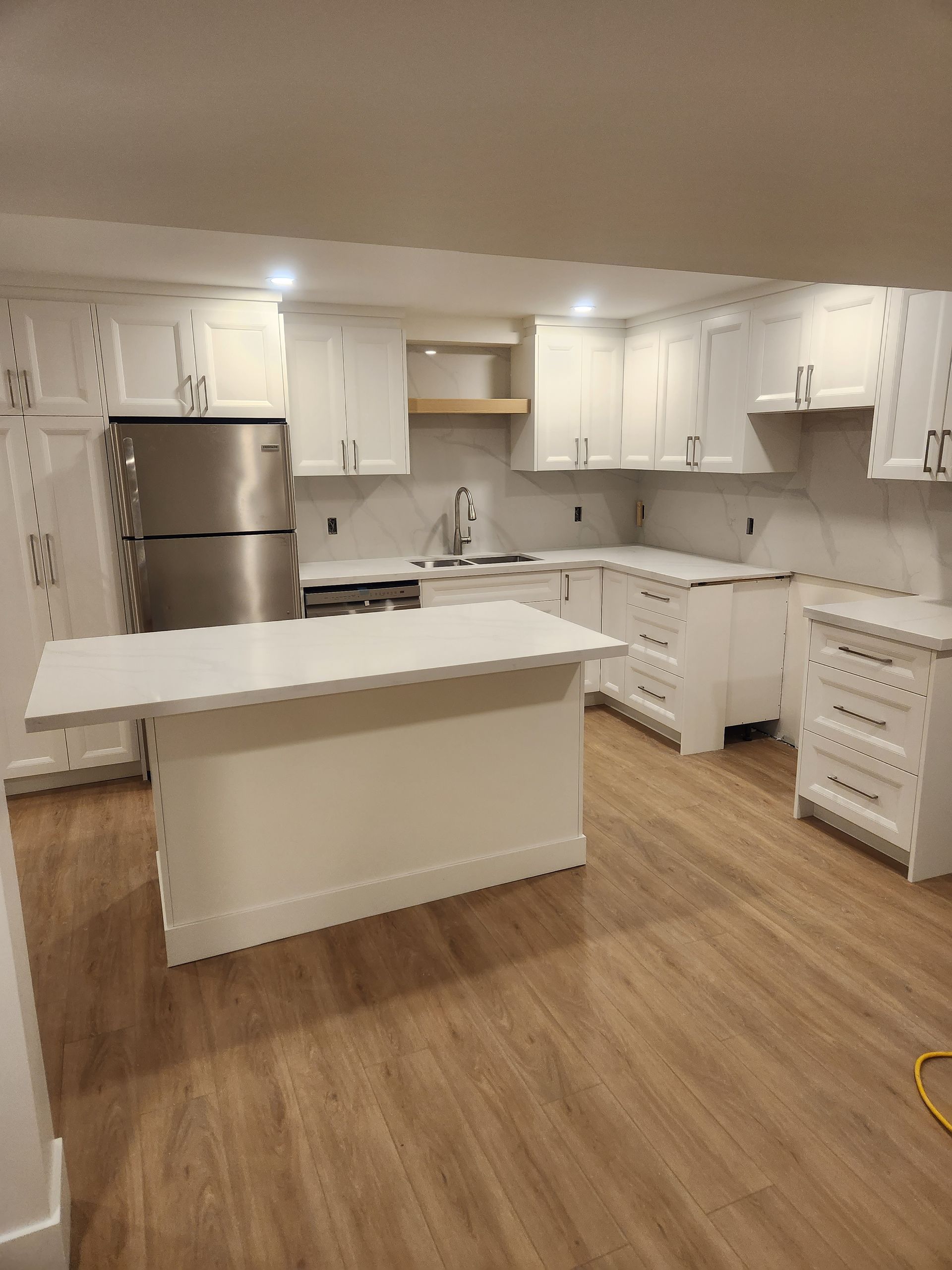 White kitchen with island, cabinets, and stainless steel refrigerator. Light wood floor.