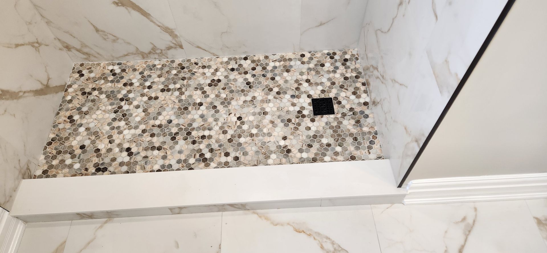 Shower with mosaic tile floor, white trim, and marble walls. Black drain.