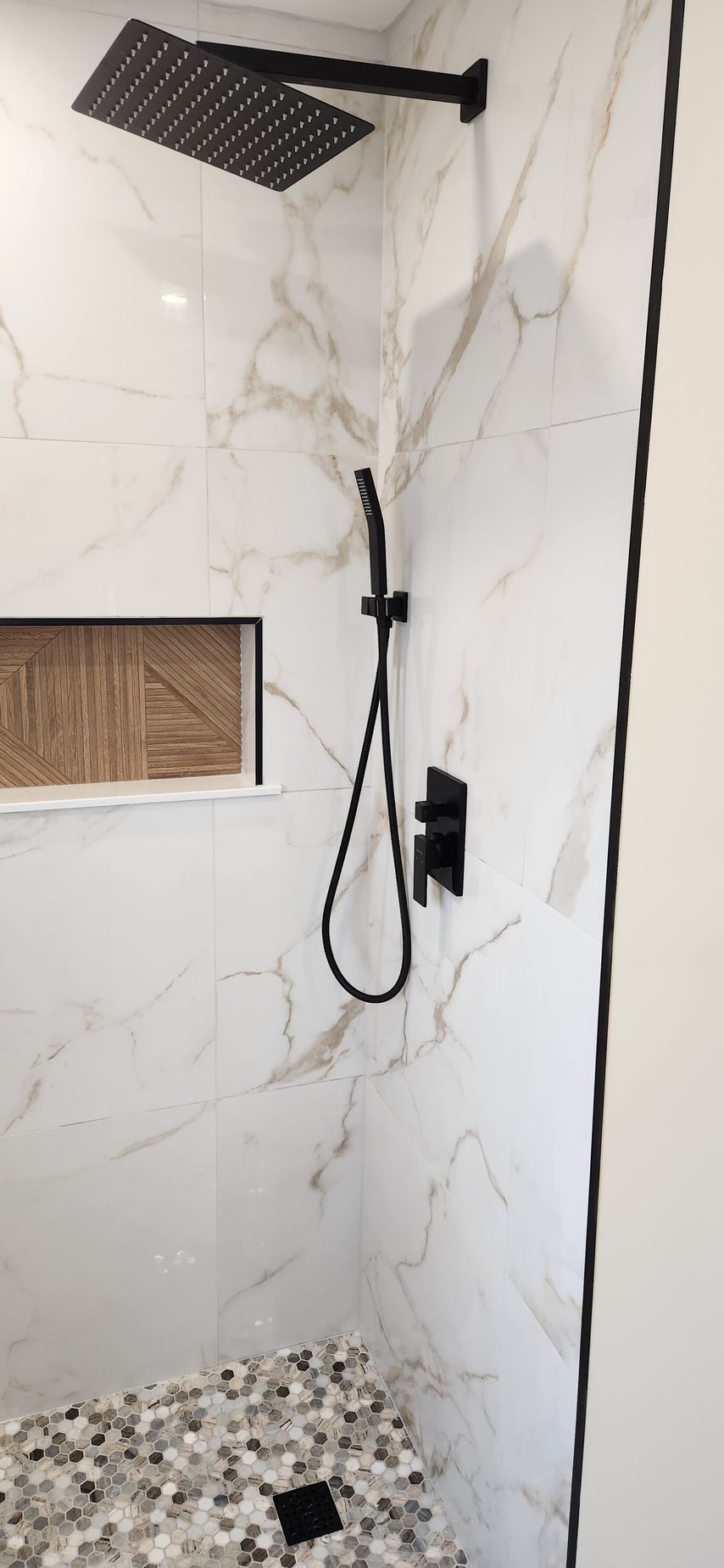 Shower with white and gold marbled tile, black fixtures, and pebble floor.