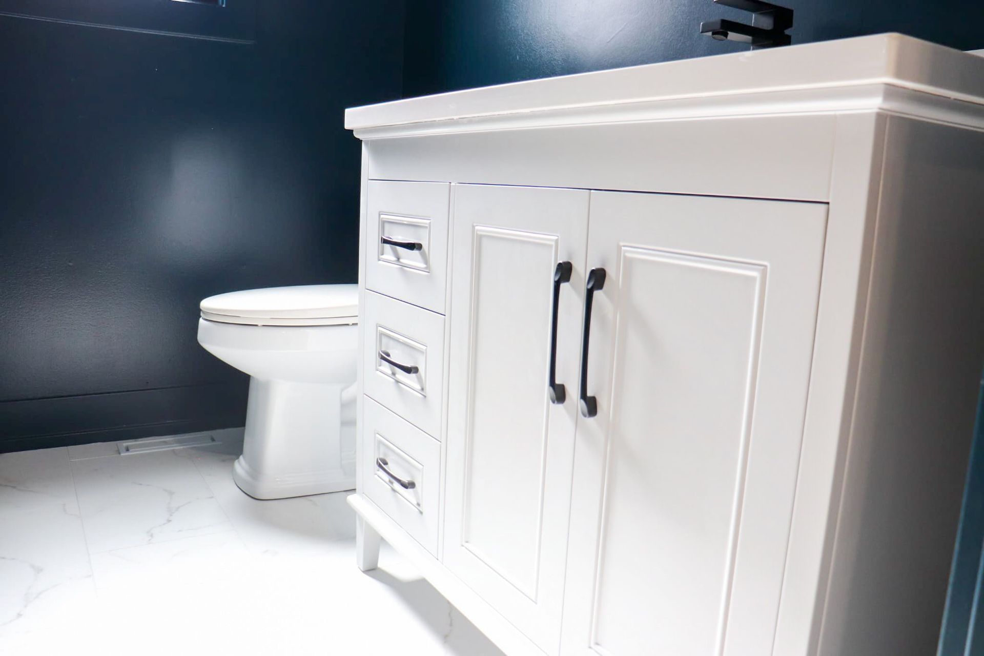 White bathroom vanity with black hardware, blue wall, toilet on white tile floor.