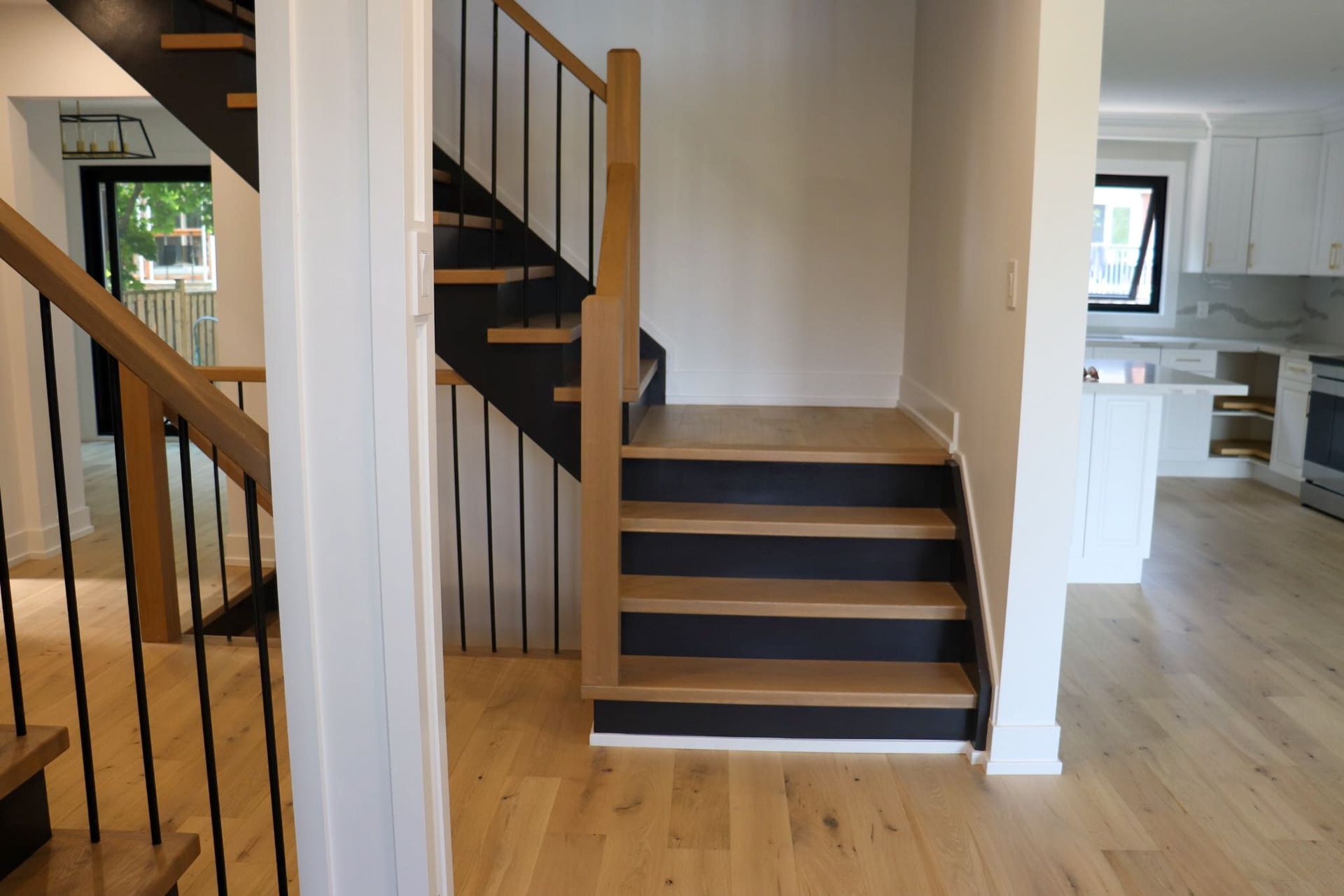 Wooden staircase with black risers, leading to a kitchen. Oak flooring, black iron railings.