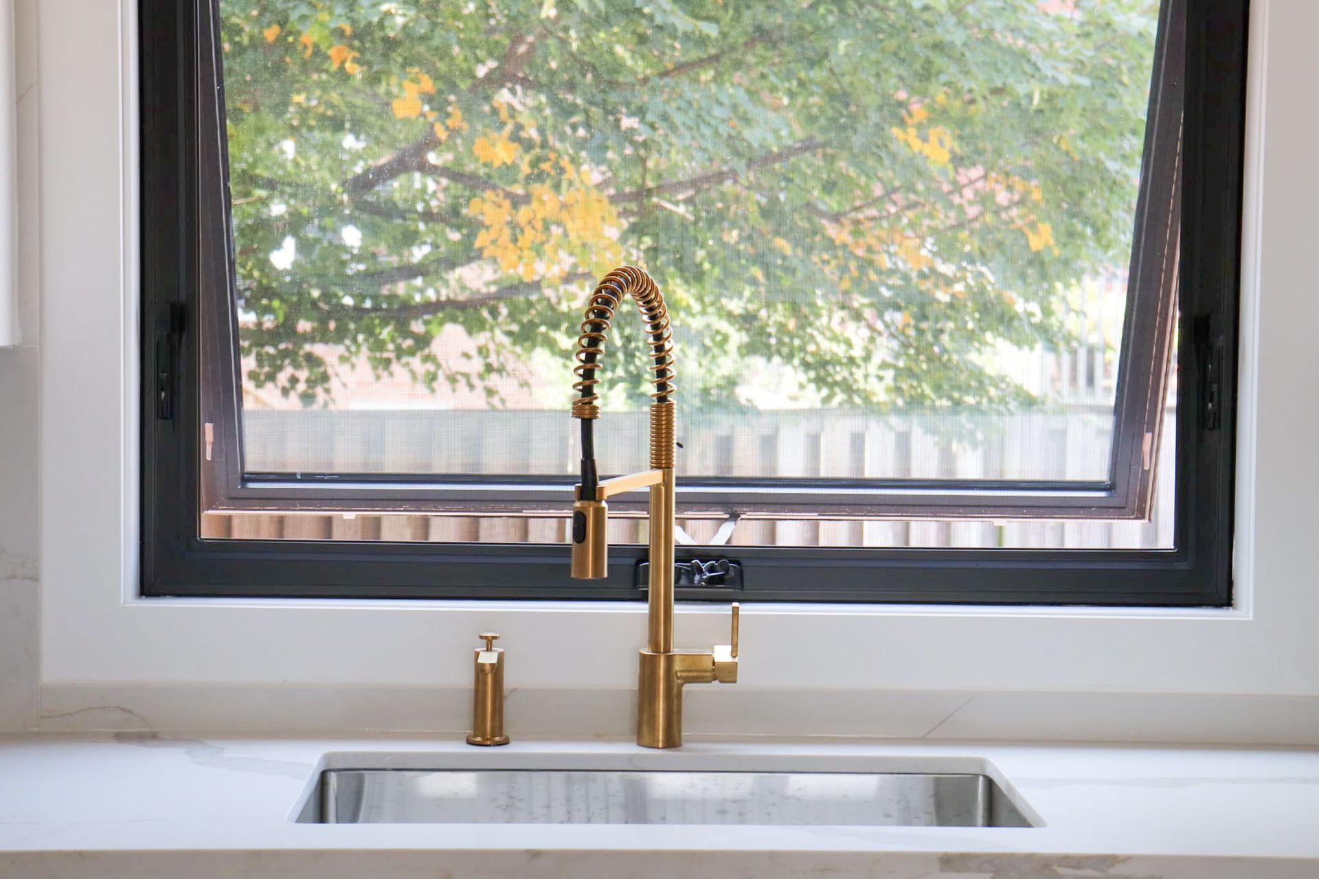 Kitchen sink with gold faucet in front of a window with a tree view.