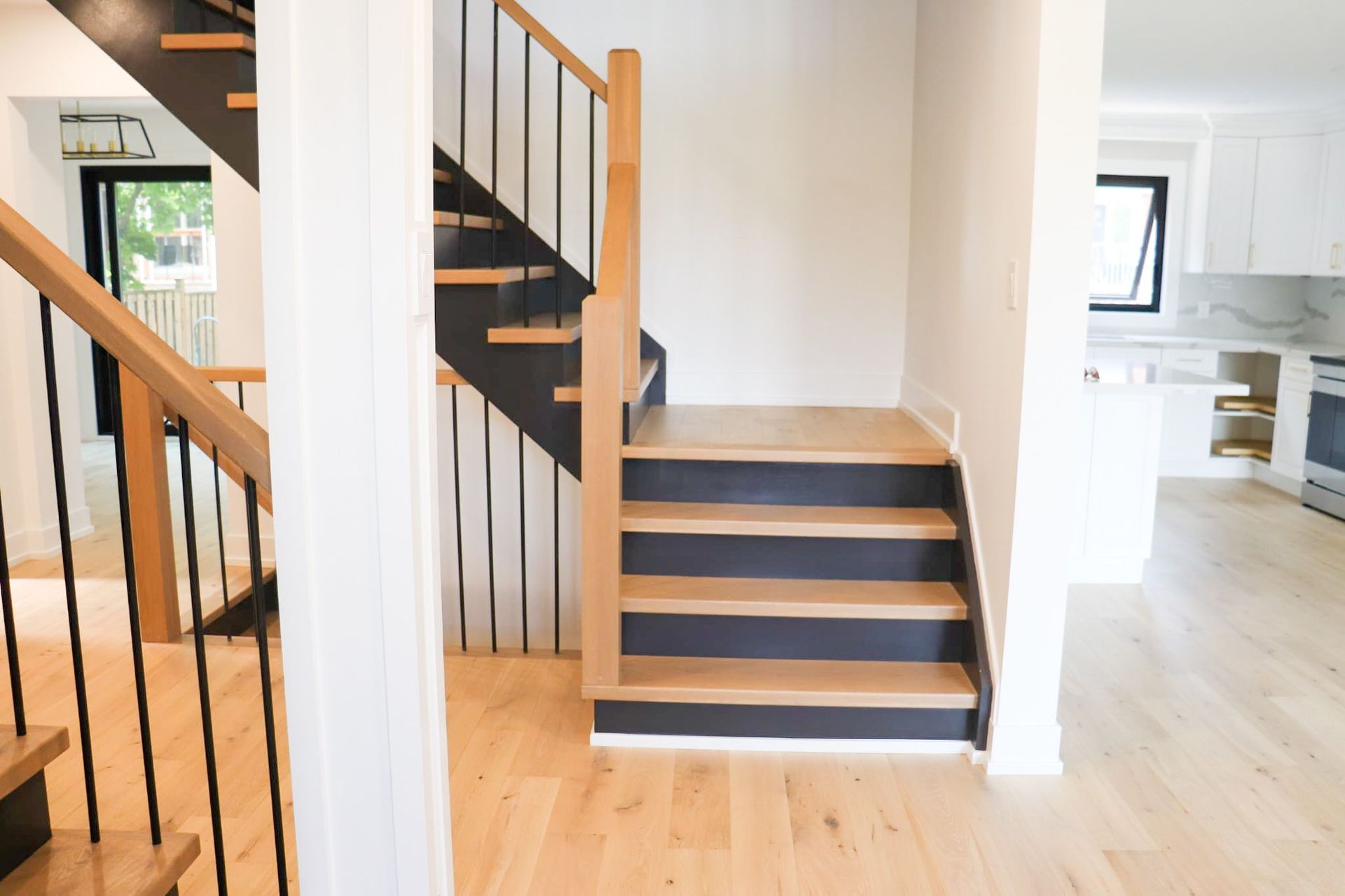 Wooden staircase with black accents and railings, leading to the upper level of a home with a kitchen visible in the background.